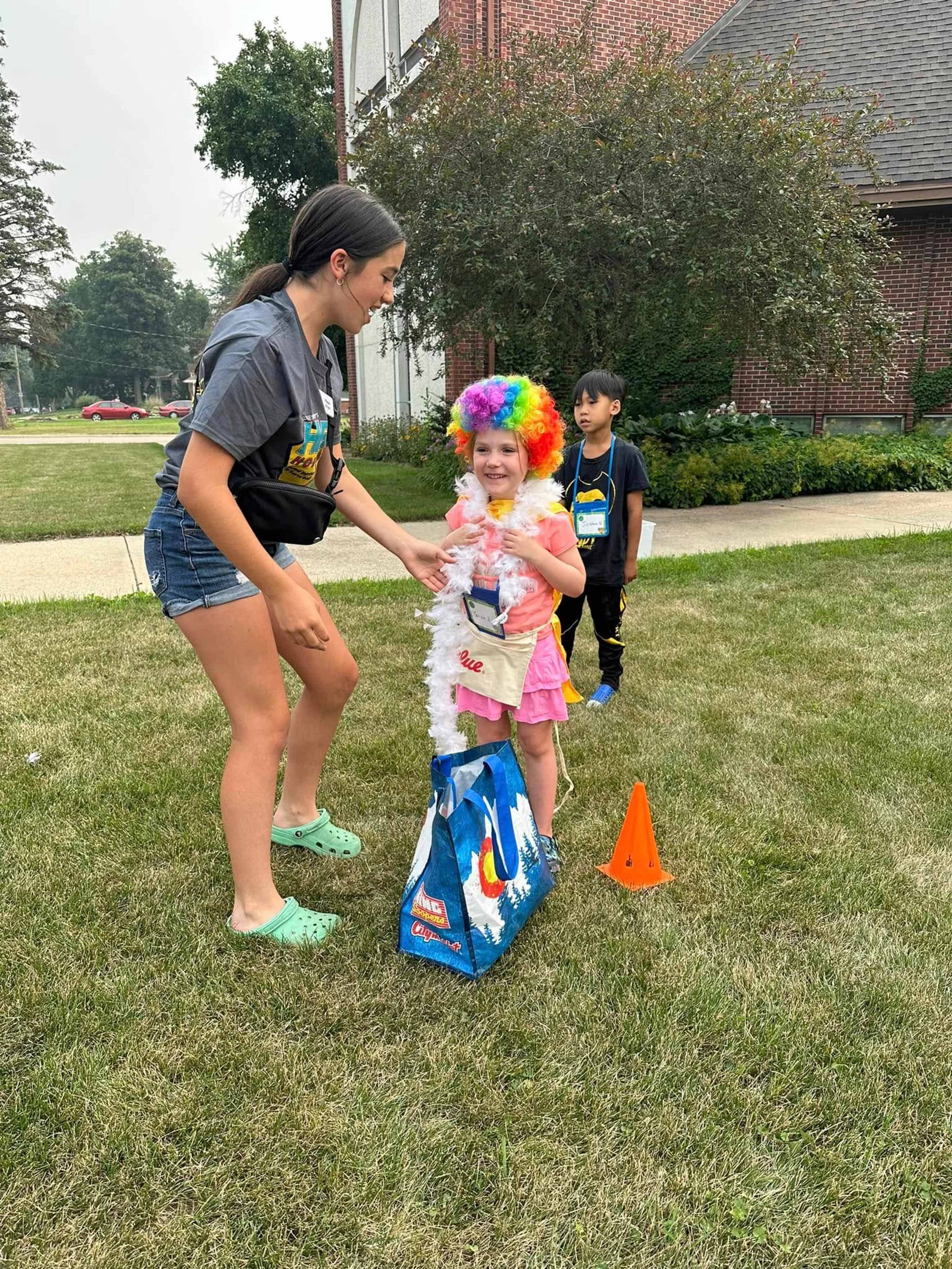 A camp counselor helps a smiling young girl in a rainbow wig and costume put something in a bag during an outdoor activity, while another boy looks on.