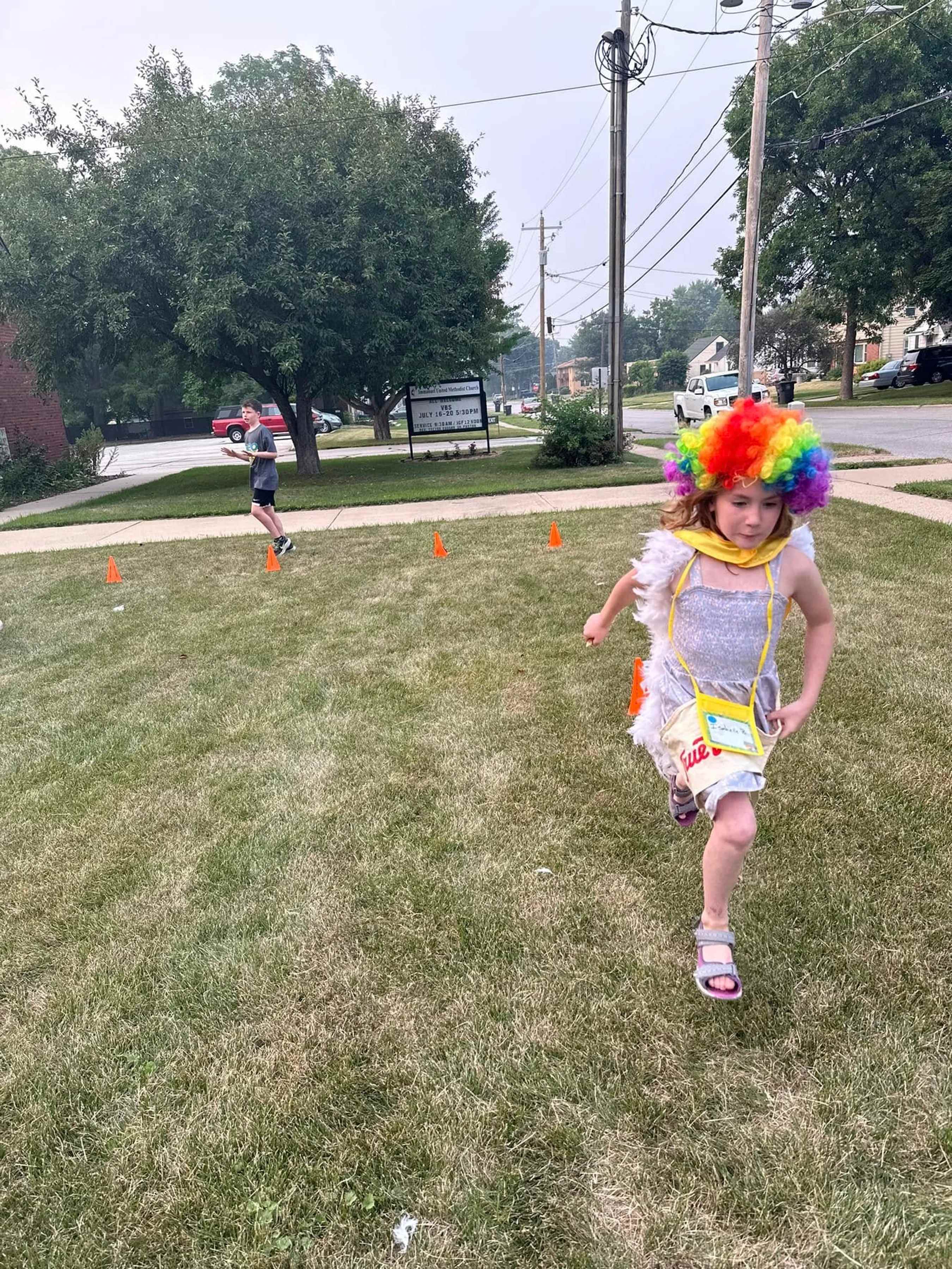 A young girl in a rainbow wig and feathered costume runs across a lawn during a race, with orange cones marking the course and a boy running behind her.
