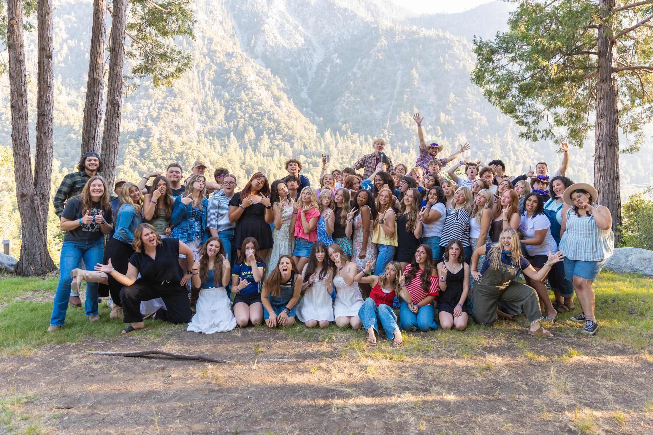 A large group of young people poses for a fun photo in a forest clearing, with a large, tree-covered mountain in the background.