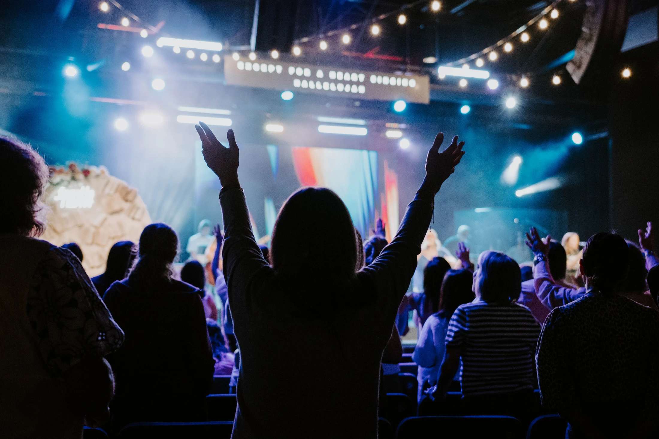 A view from behind a crowd at a church service, with one person in the center raising their hands in worship towards a brightly lit stage.