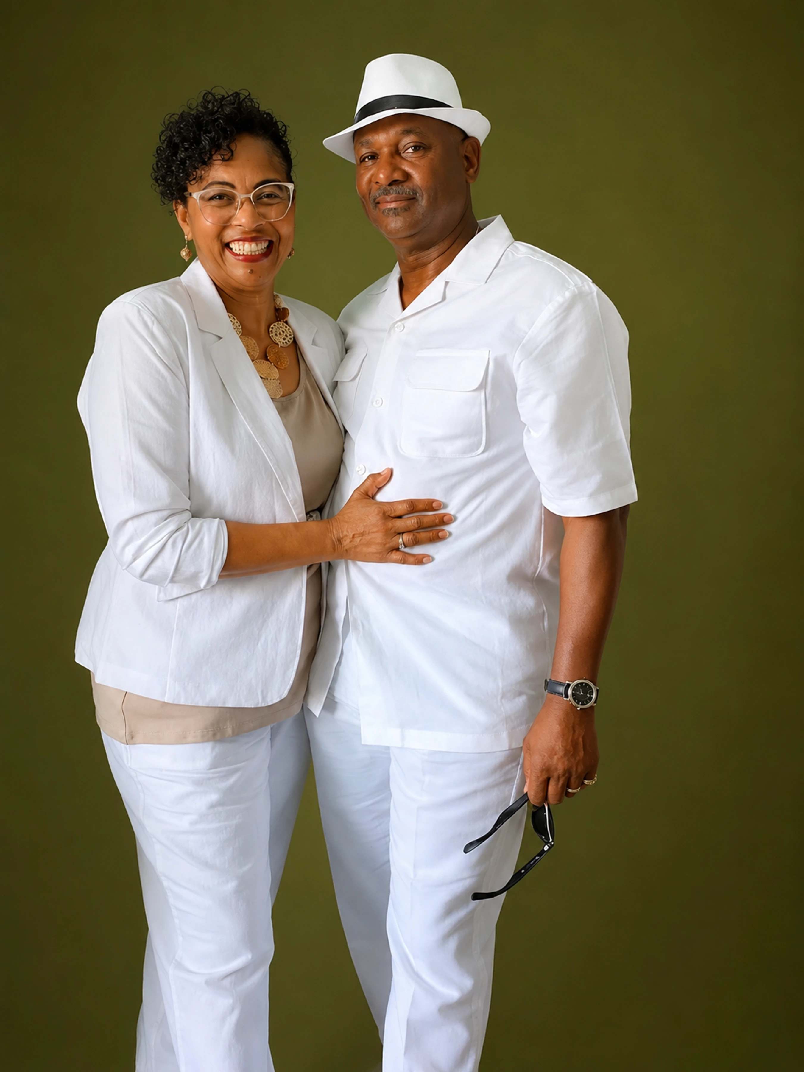 A stylish, smiling Black couple poses in matching white outfits against an olive green background. The man wears a fedora and the woman wears glasses.
