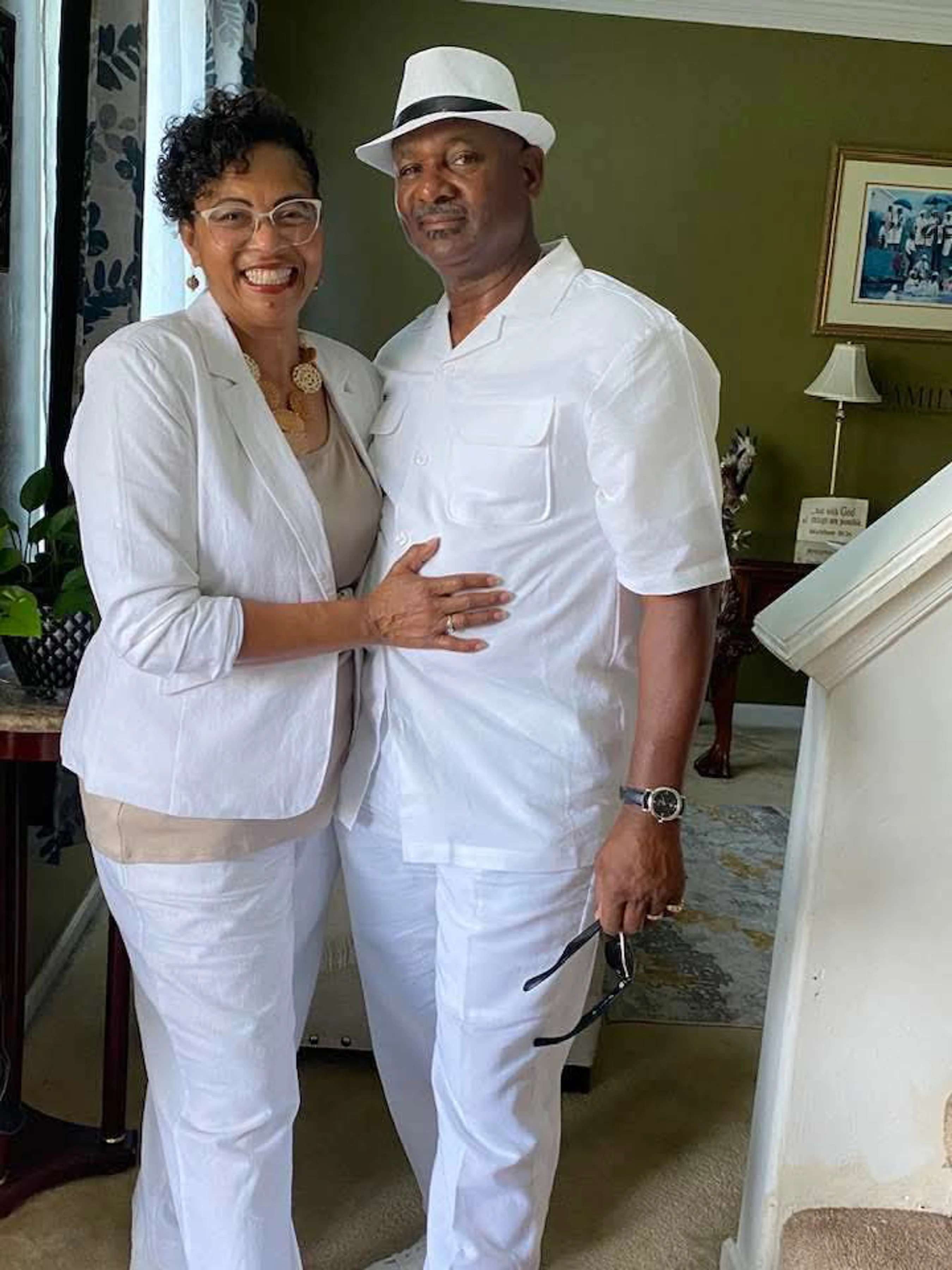A smiling Black man and woman pose together indoors, both wearing all-white outfits. The man wears a fedora, and the woman has her arm around his waist.
