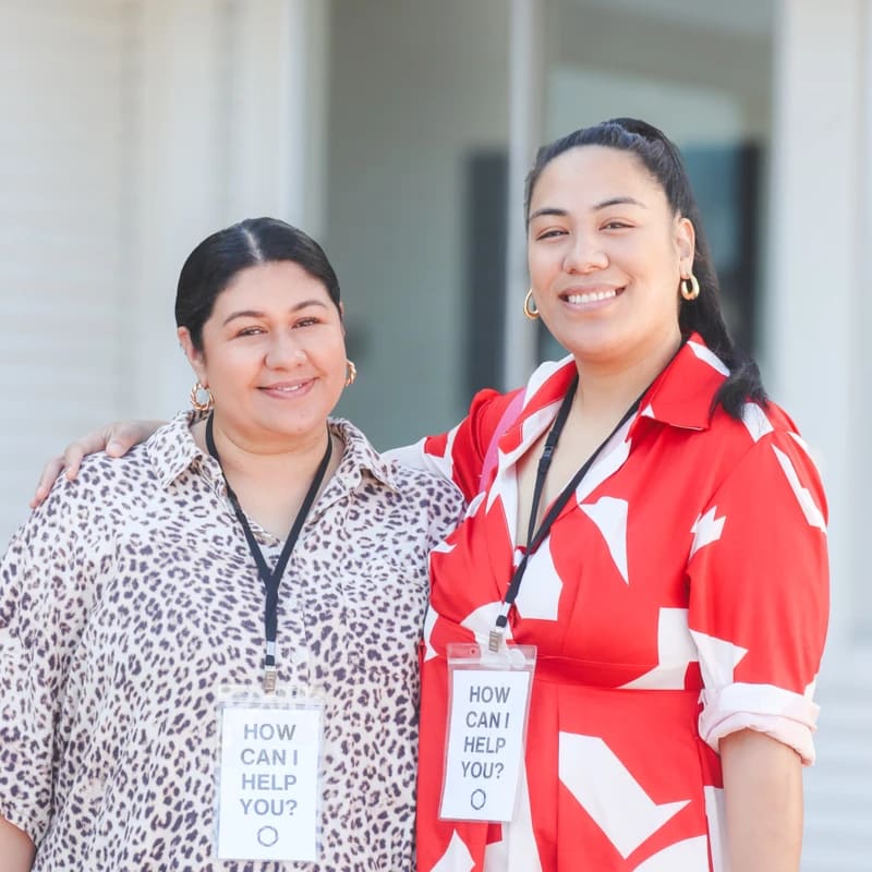 Two women stand side-by-side outdoors, smiling at the camera. They each wear a lanyard with a badge that asks, 'How can I help you?'.