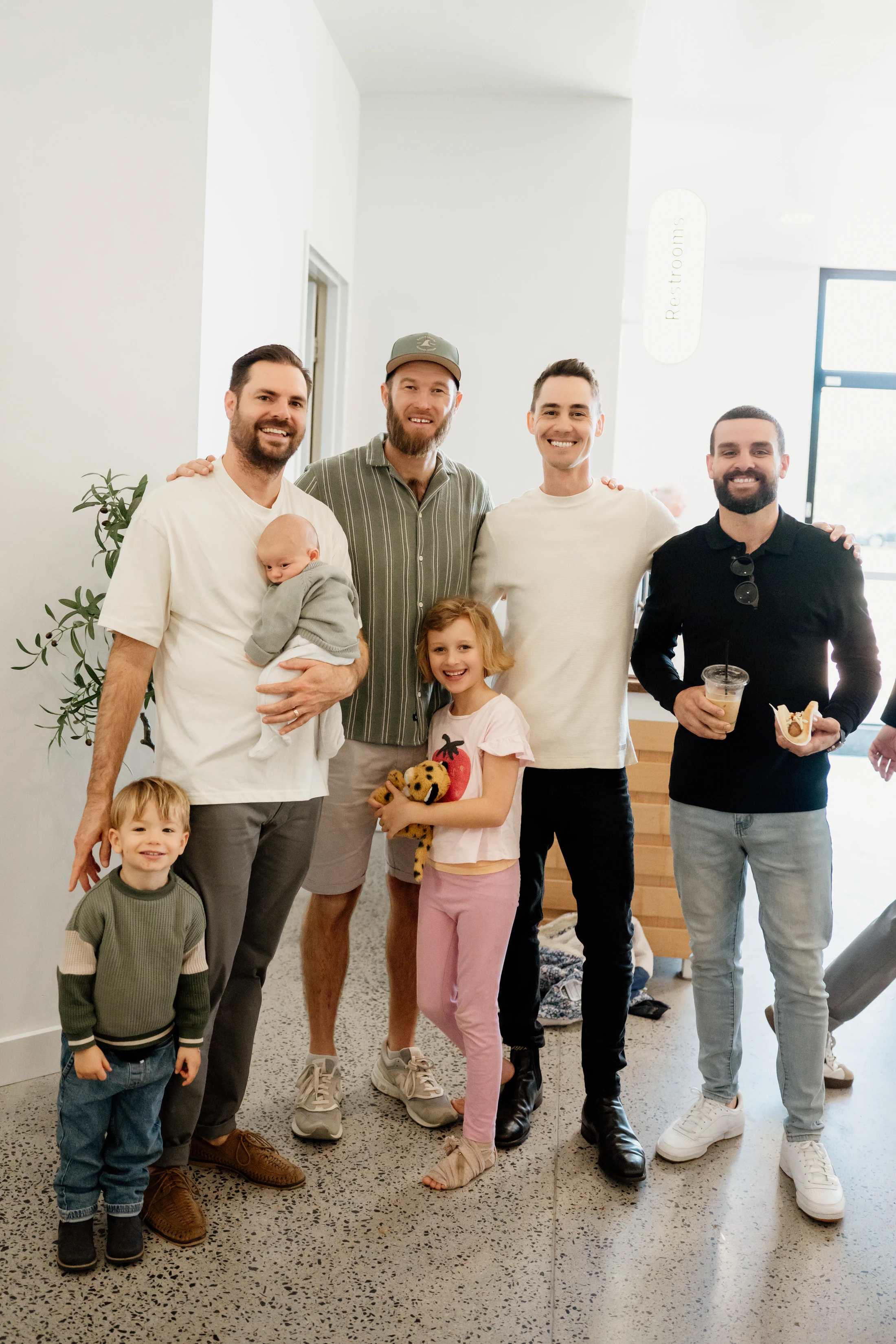 A group of four men and three young children stand together smiling for a photo indoors. One man holds a baby, and a girl holds a stuffed animal.