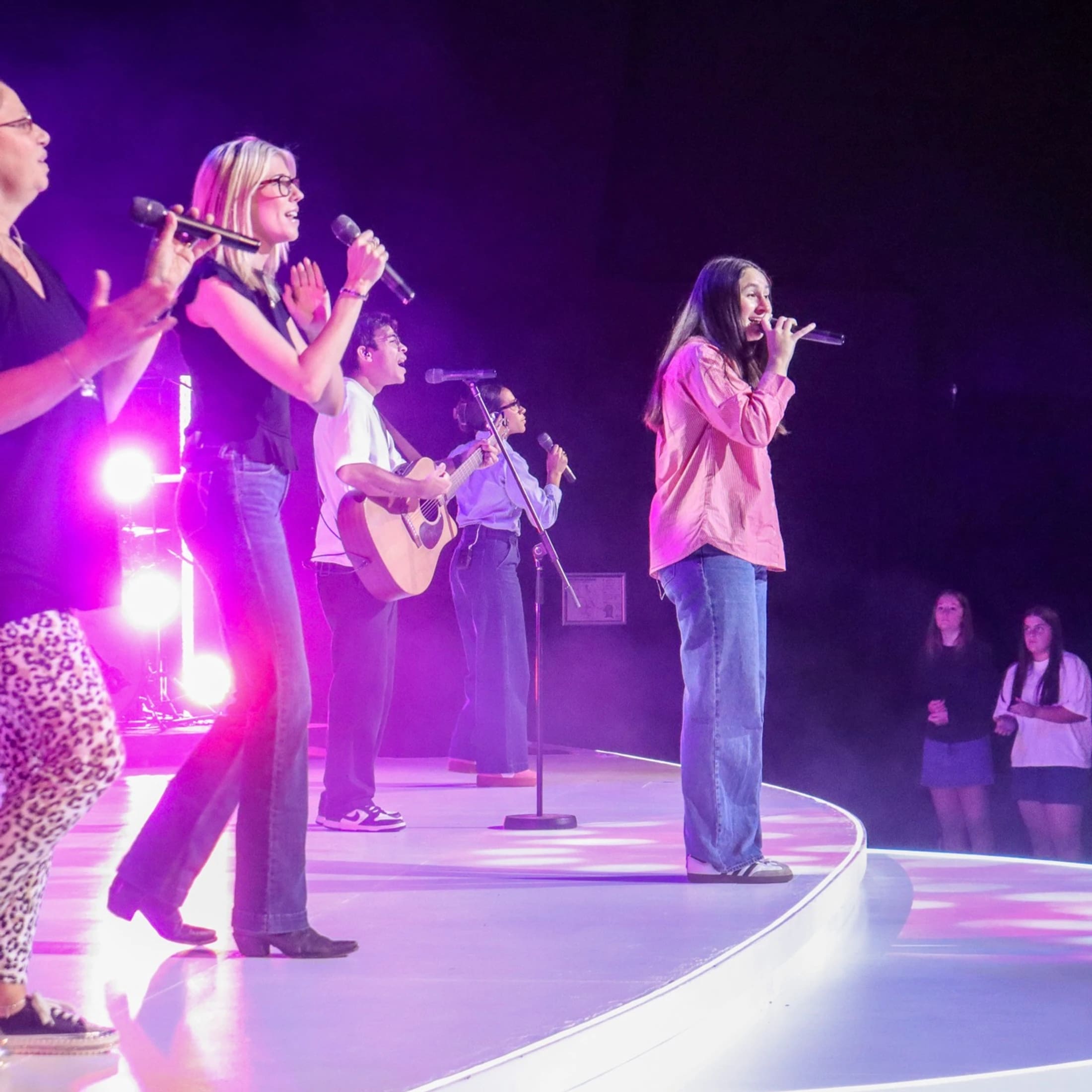 A diverse group of people sings and plays guitar on a circular stage, illuminated by vibrant purple and pink lights during a live performance.