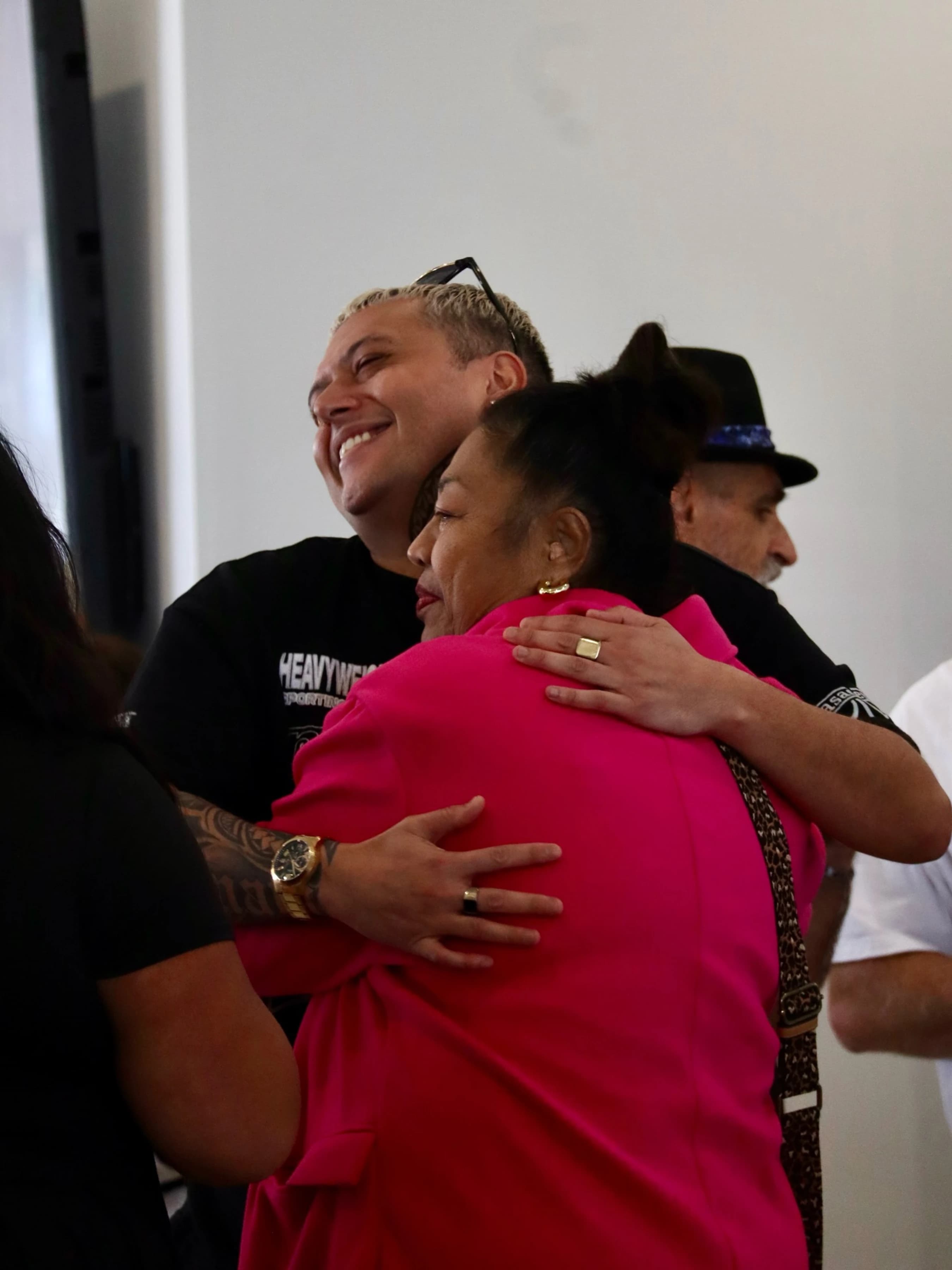 A smiling man with short blonde hair and arm tattoos warmly hugs a woman in a bright pink jacket. Another person in a fedora is visible in the background.