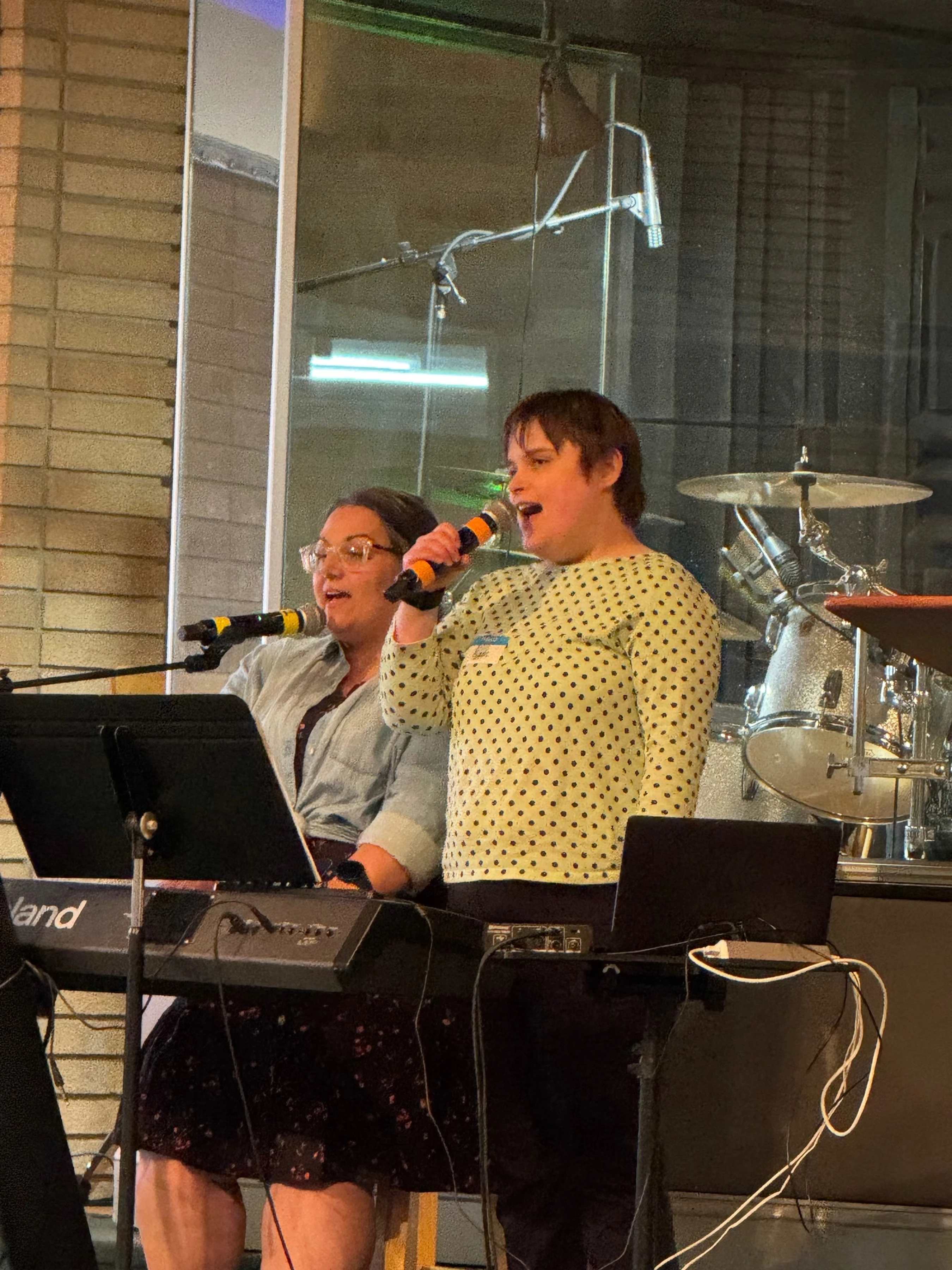 Two women sing into microphones on stage. One stands in a polka dot shirt, and the other sits at a keyboard. A drum set is visible in the background.