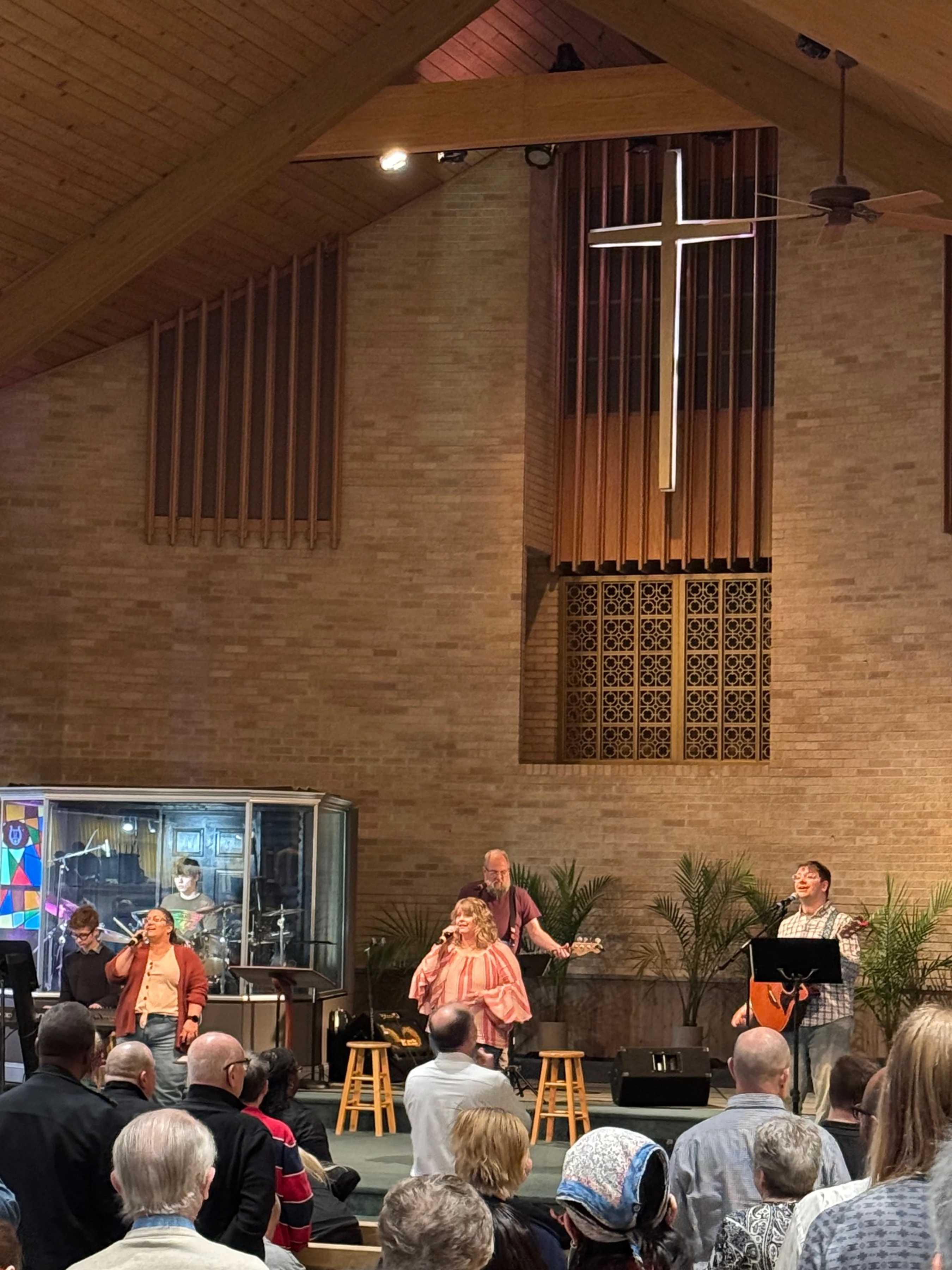 A band with singers and musicians performs on a stage for a congregation inside a church with high wood-paneled ceilings and an illuminated cross.