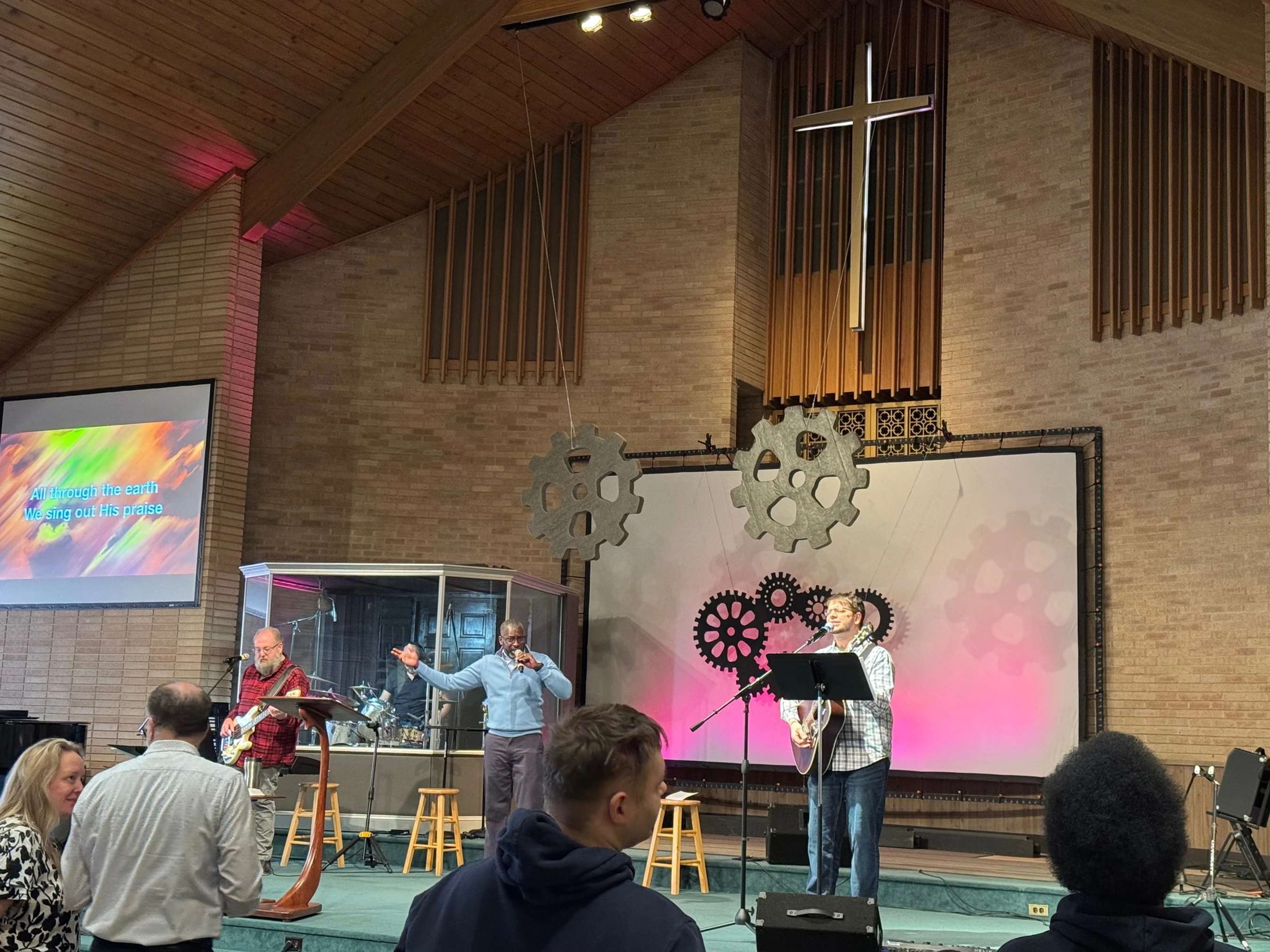 A worship band with several musicians performs on a stage for a congregation inside a church with a large, illuminated cross hanging in the background.