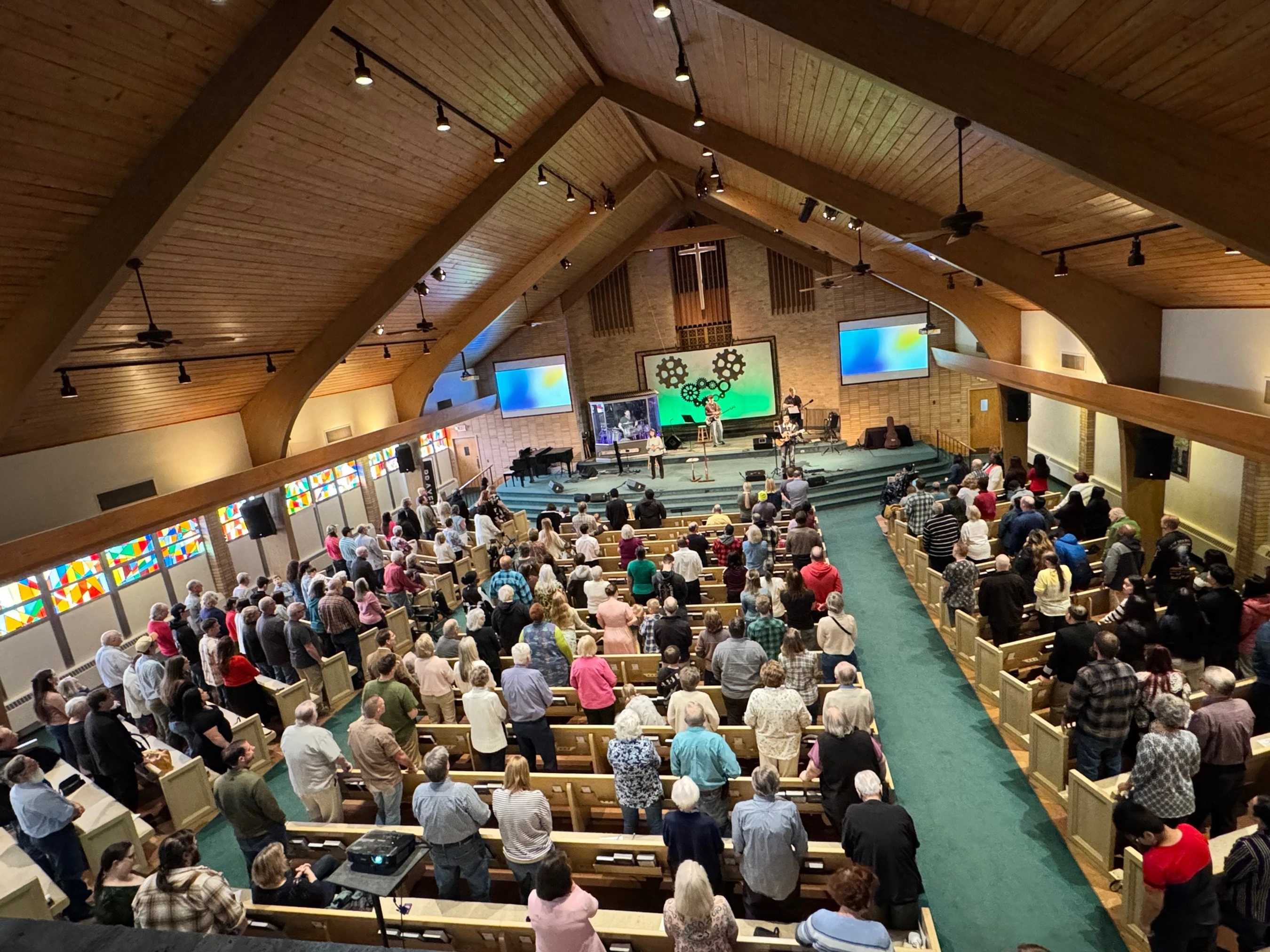 A high-angle view of a large congregation standing in pews during a contemporary church service, with a band playing on stage under a vaulted wooden ceiling.