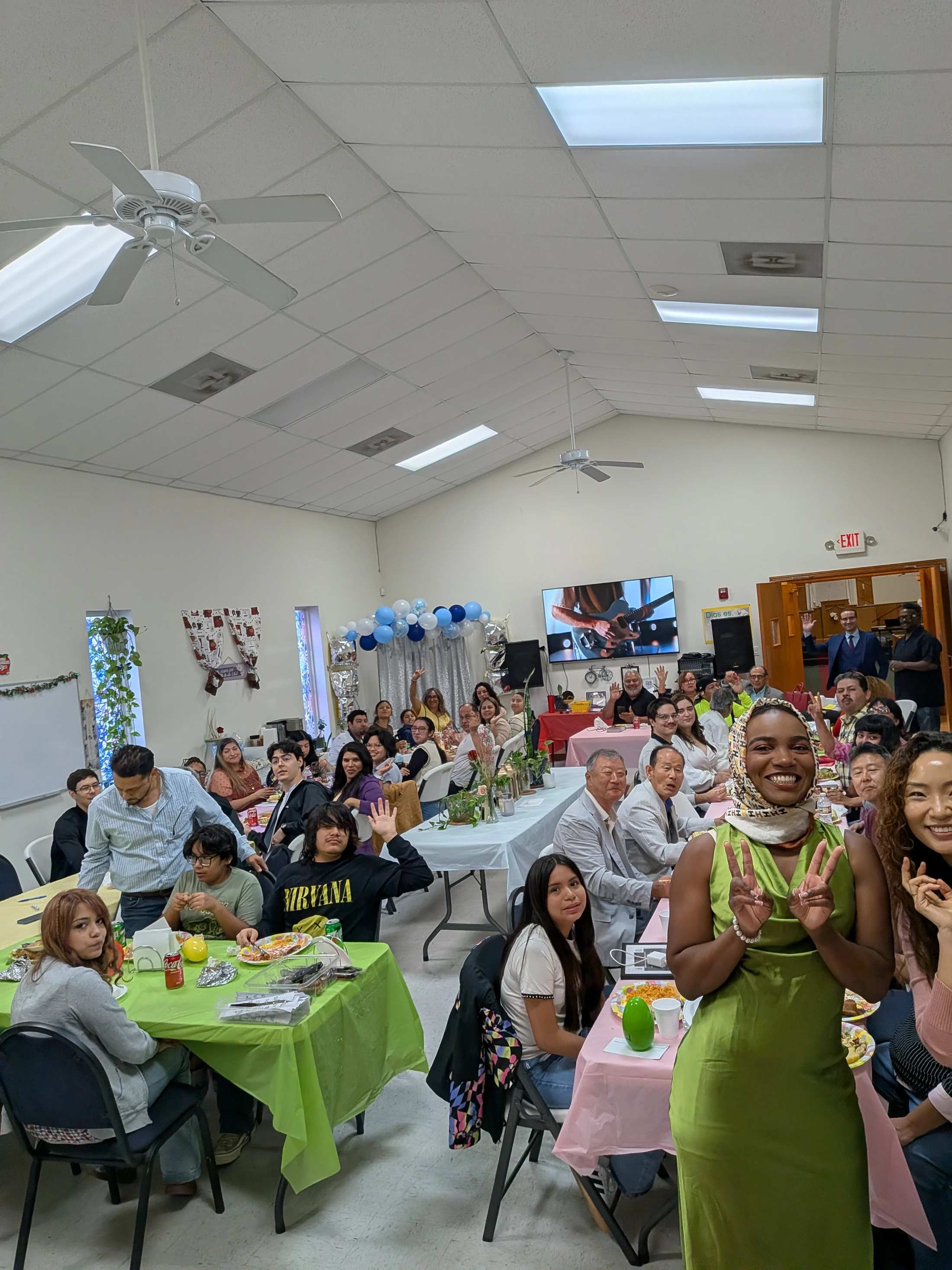A large, diverse group of people at an indoor party. They are sitting at long tables, eating and smiling for the camera. A woman in front wears a green dress.