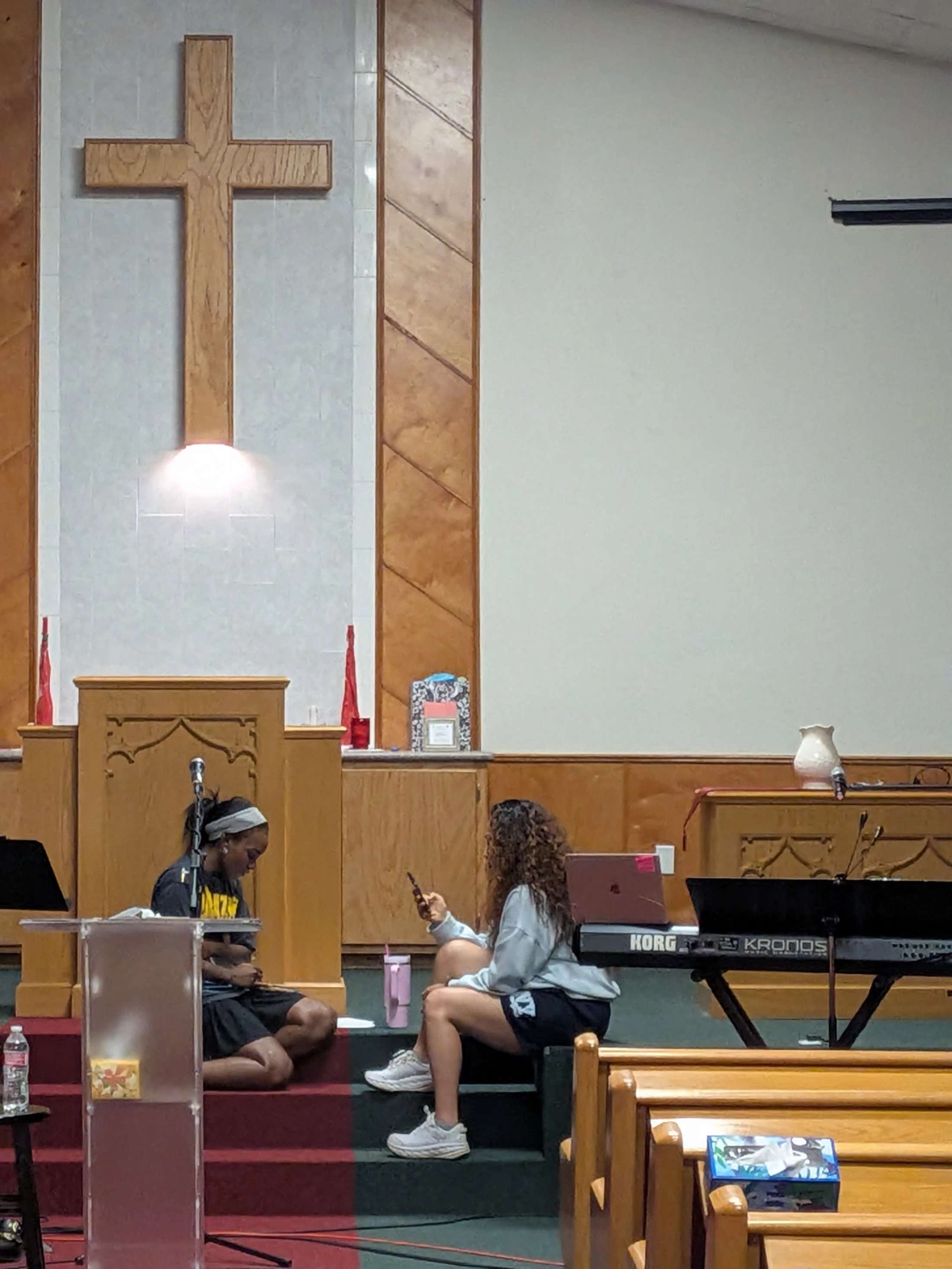 Two young women sit casually on the steps of a church altar. A large wooden cross is mounted on the wall behind them. One woman looks at her phone.