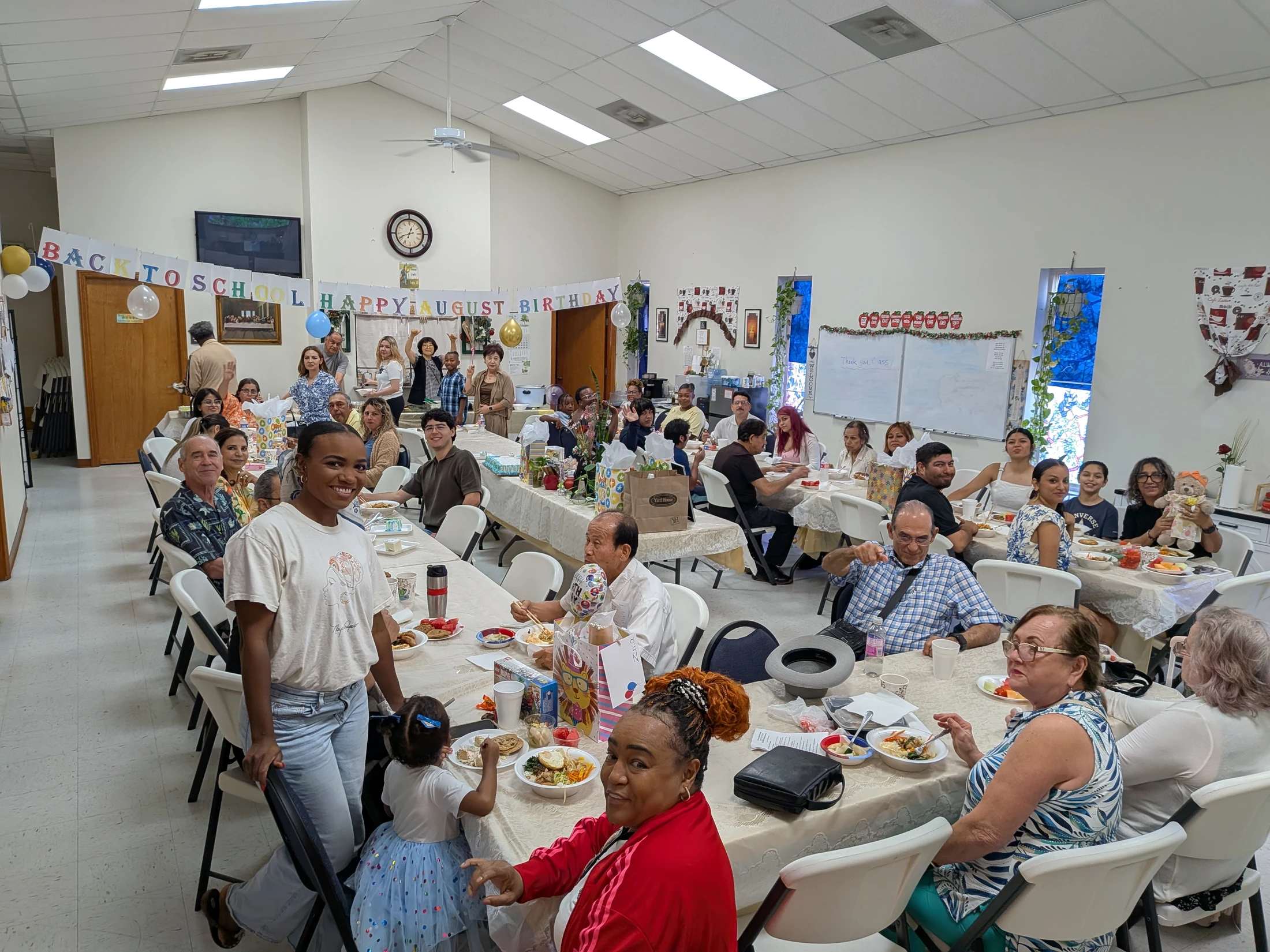 A diverse group of people of all ages enjoying food and socializing at long tables in a decorated hall for a celebration.