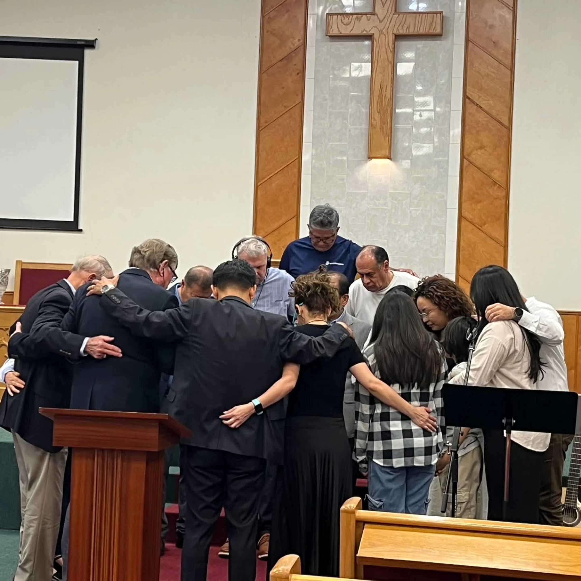 A diverse group of people stand in a huddle at the front of a church, praying together with heads bowed and arms around each other's shoulders.