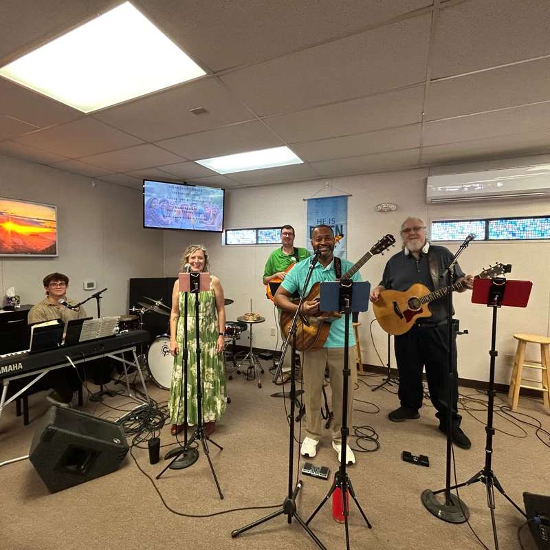 A smiling five-person worship band plays music with guitars and a keyboard on a small stage in a church, surrounded by sound equipment.