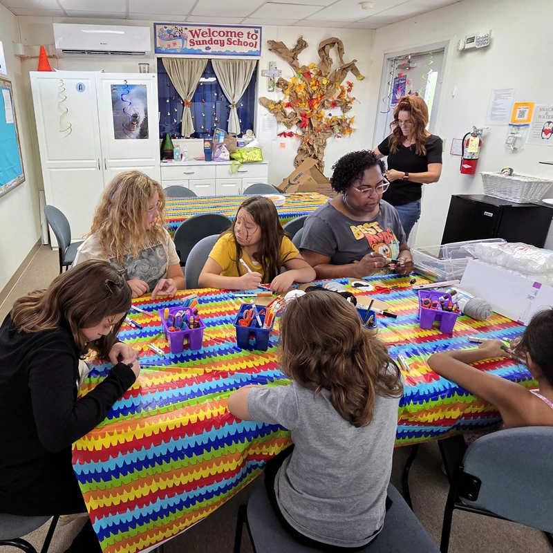 A group of children and adults sit at a long table with a colorful tablecloth, engaged in an arts and crafts project in a Sunday school classroom.