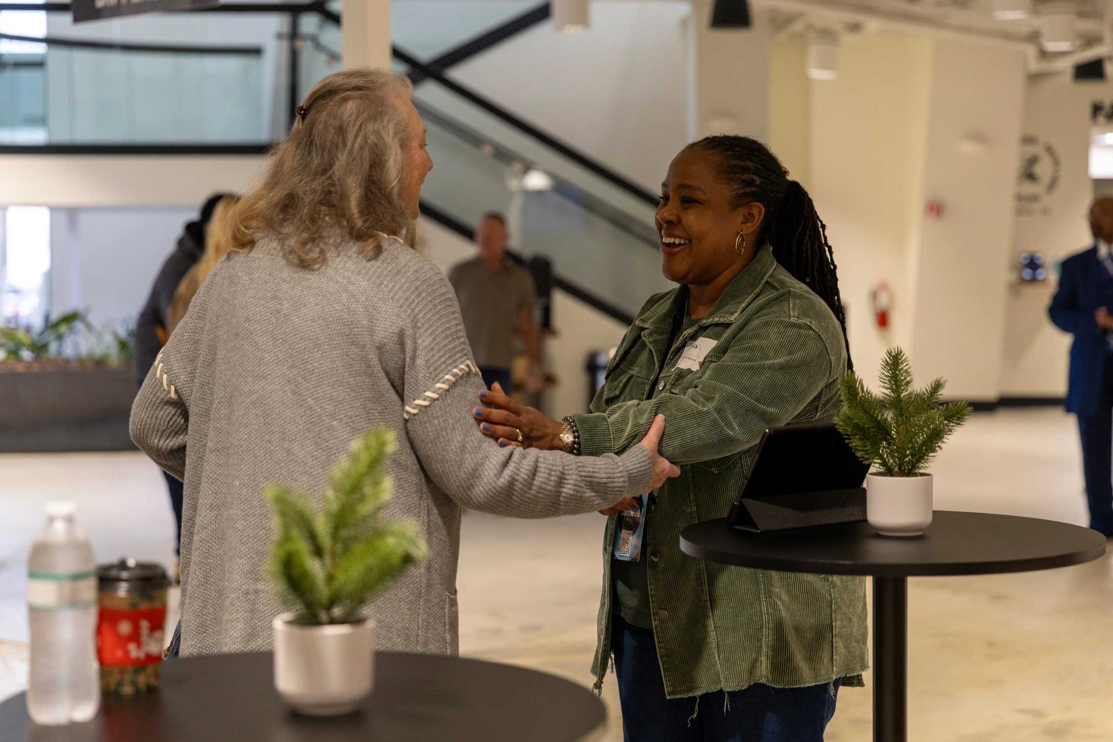 Two women stand facing each other, holding arms and smiling. One has long gray hair and a gray sweater; the other has locs and a green corduroy jacket.