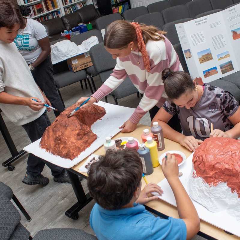 A group of students works together around a table, painting 3D paper-mache models of mountains and volcanoes in a classroom setting with bookshelves.