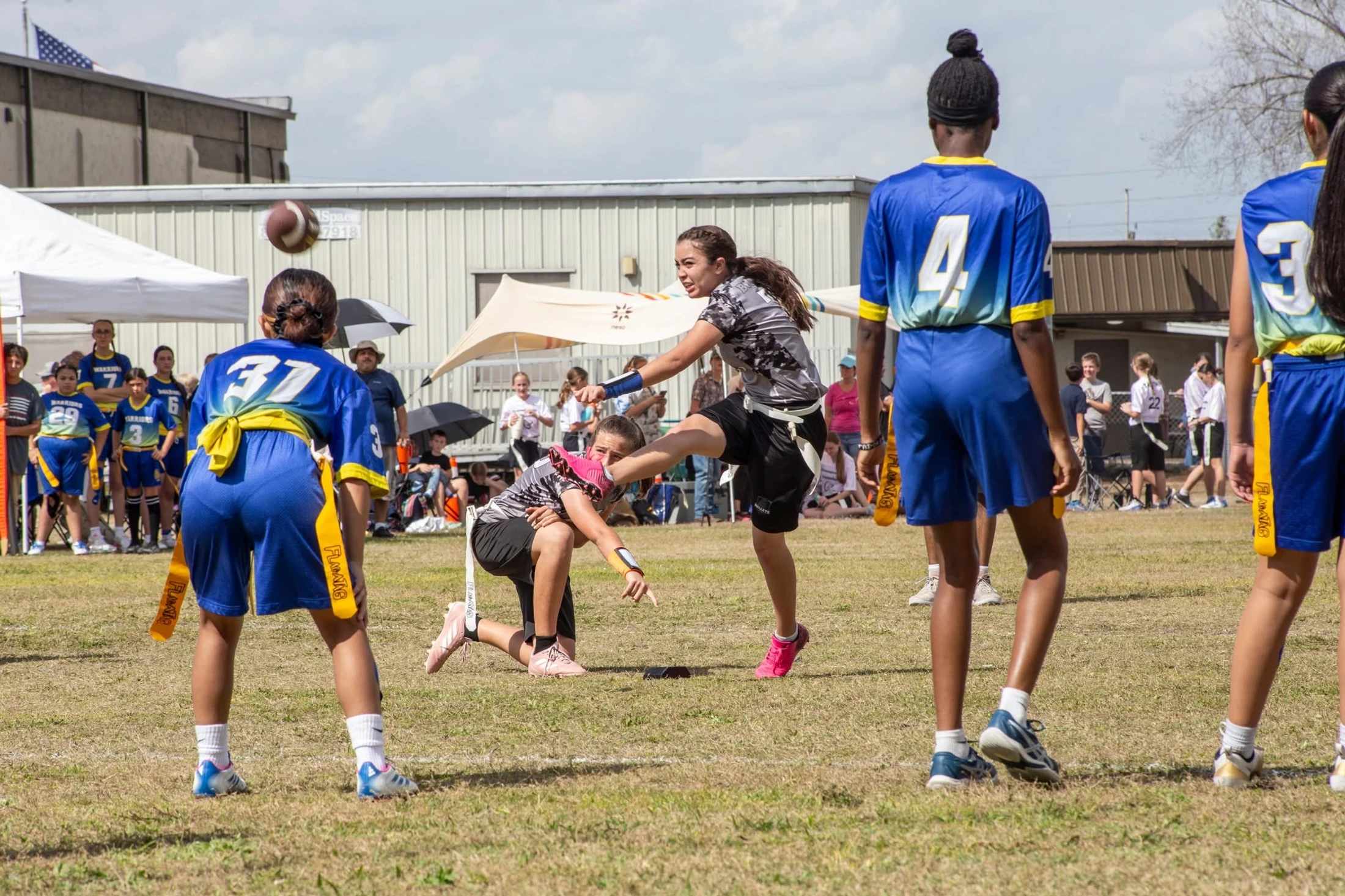 A group of young girls playing flag football on a grassy field. One girl in a grey uniform throws the ball as players from the opposing blue team watch.