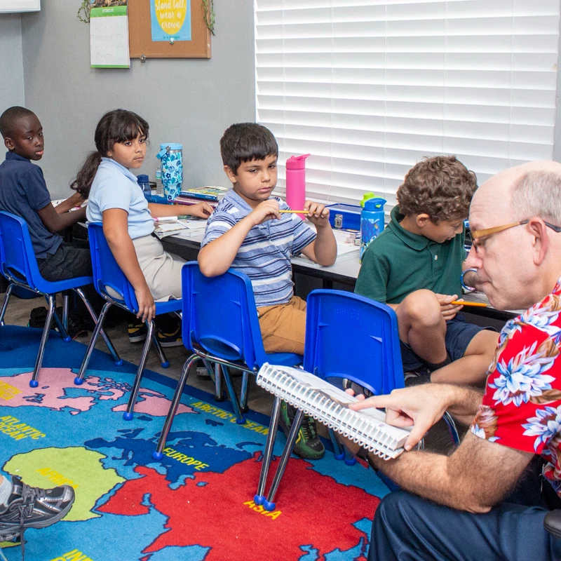 A male teacher in a red floral shirt sits on the floor, showing a book to a group of diverse elementary school students sitting at their desks.