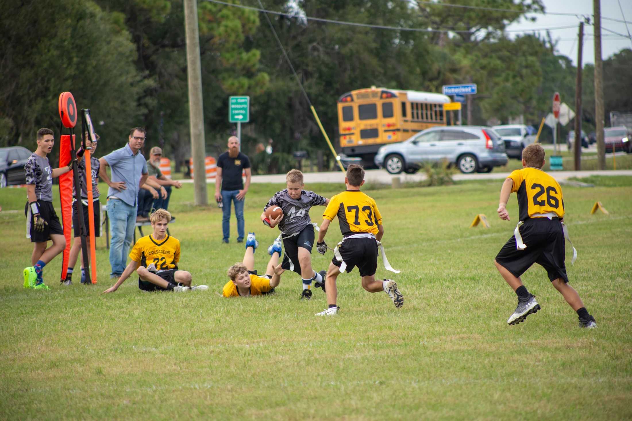 A young boy in a grey jersey runs with a football during a flag football game, as other boys in yellow jerseys try to stop him on a green field.