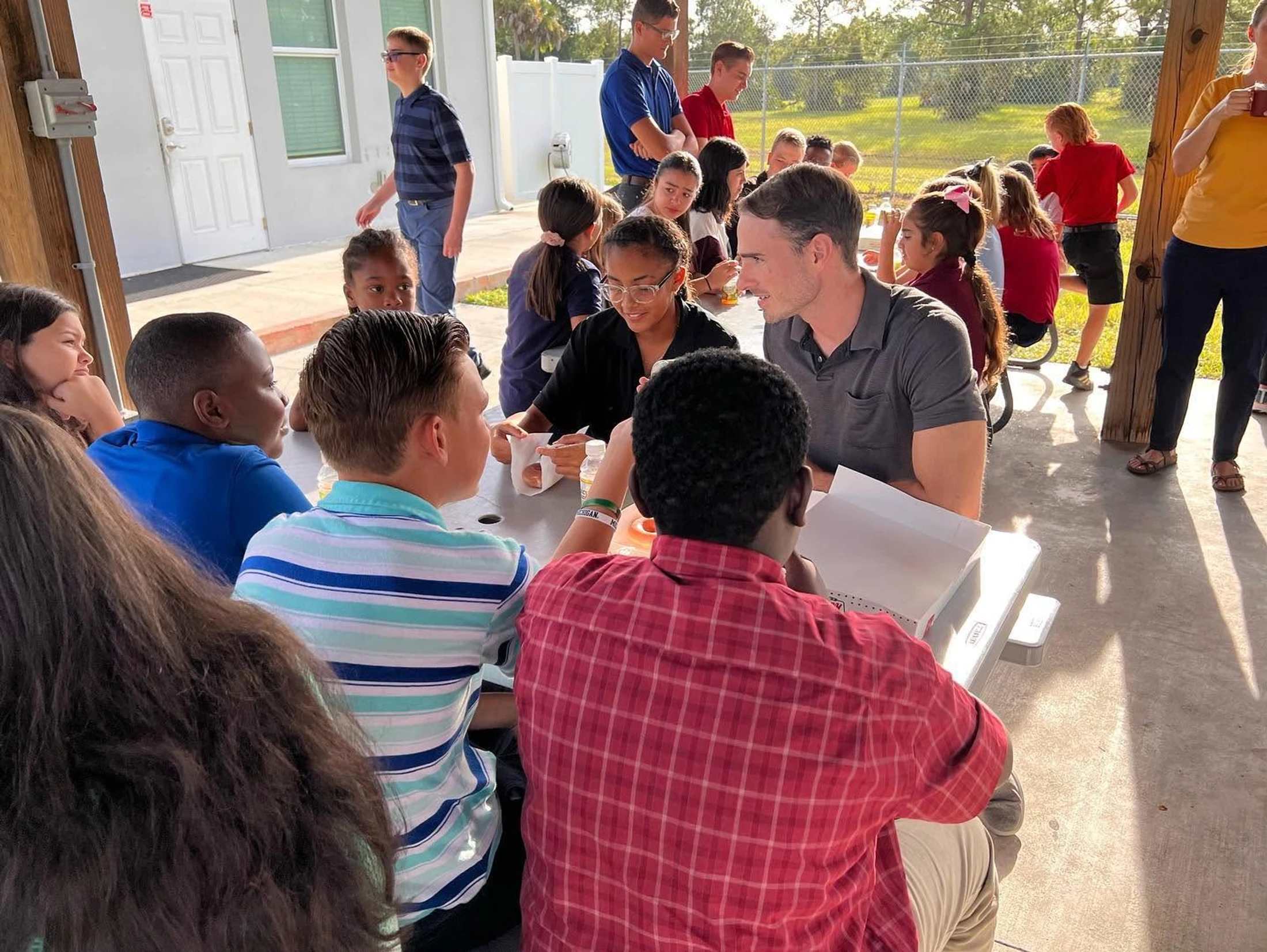 A group of diverse children and a few adults are gathered around a picnic table in a covered outdoor area, engaged in conversation on a sunny day.