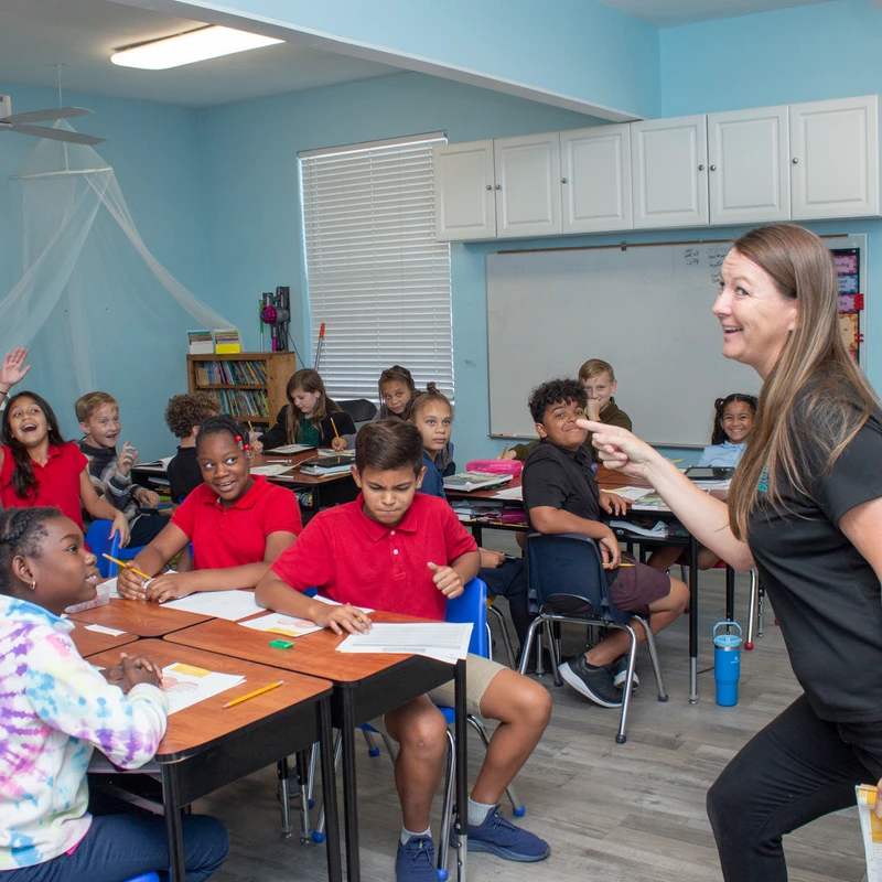 A female teacher smiles and points at a student in a lively classroom. Diverse students sit at their desks, many smiling and participating in the lesson.