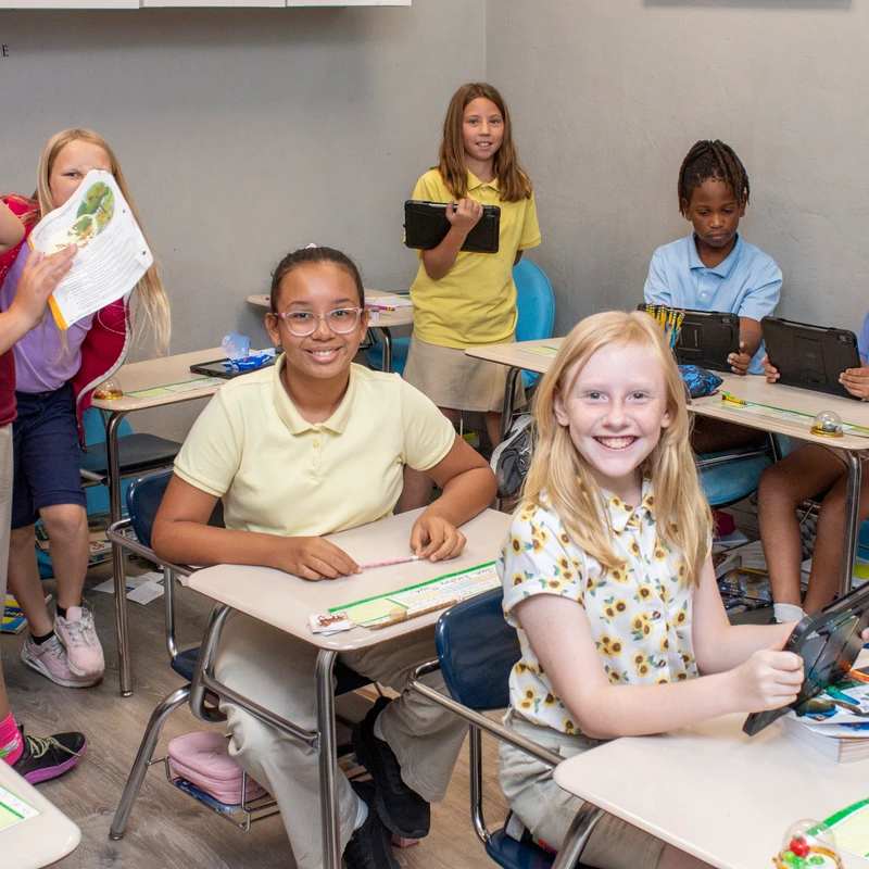 A diverse group of young students in a classroom. Two girls in the foreground smile at the camera while classmates work at their desks with books and tablets.