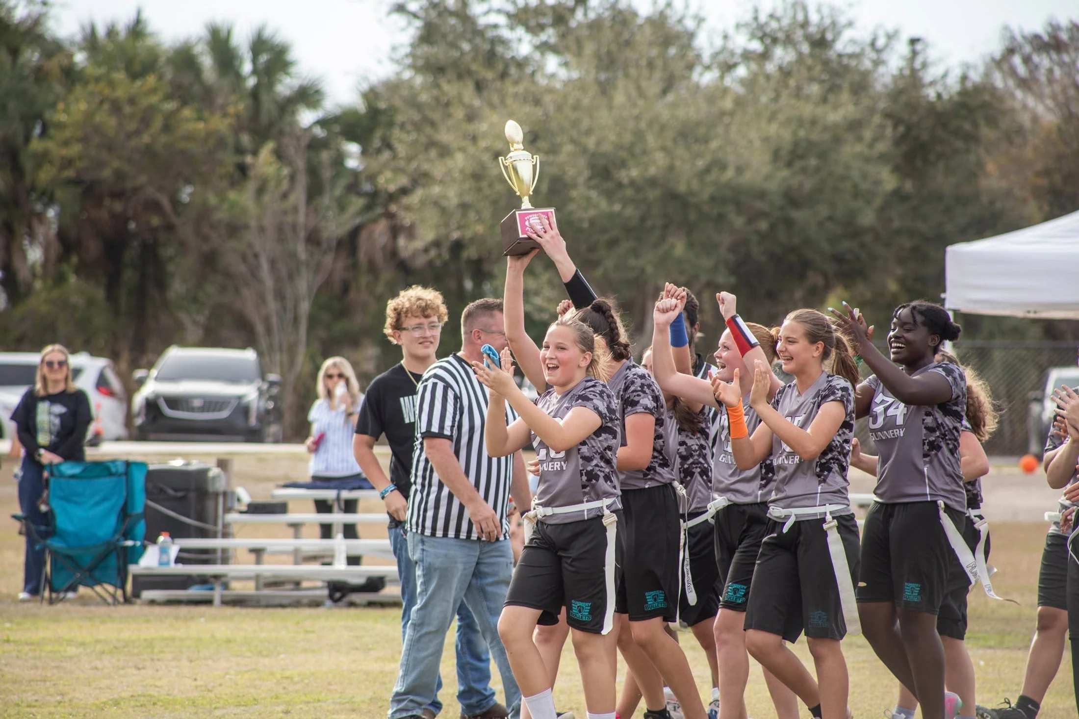 A youth girls' flag football team in gray and black uniforms joyfully cheers on a grassy field, with one player holding a large gold trophy high in the air.