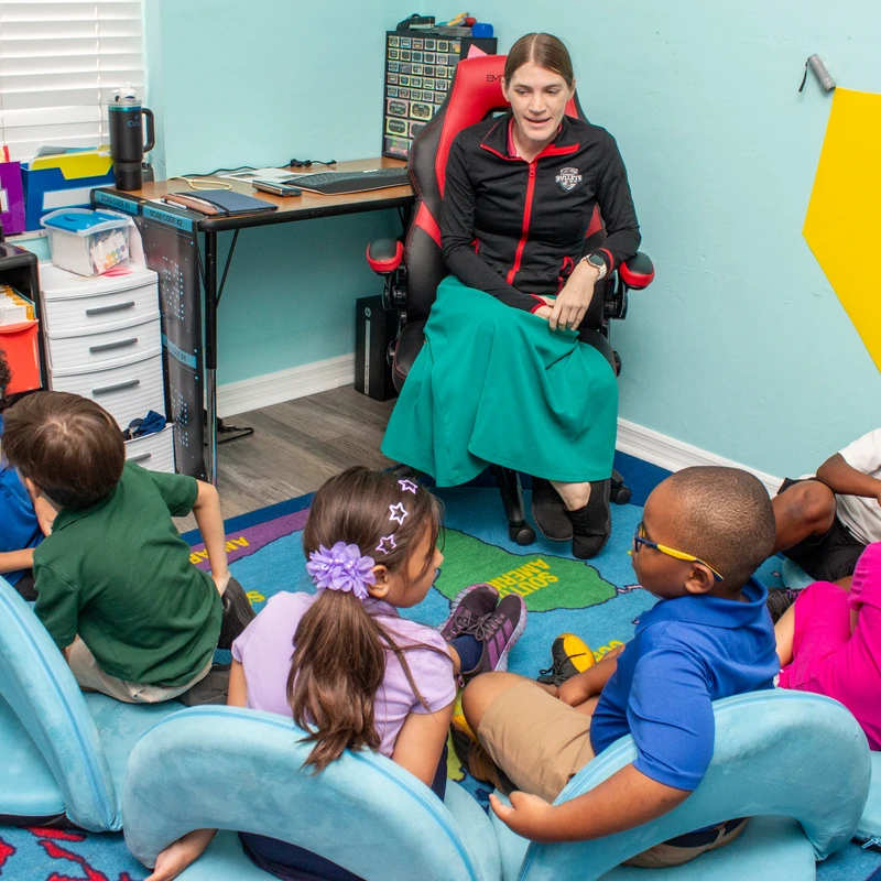 A teacher sits in a chair facing a group of diverse young students who are sitting in a semi-circle on blue floor chairs on a colorful rug.