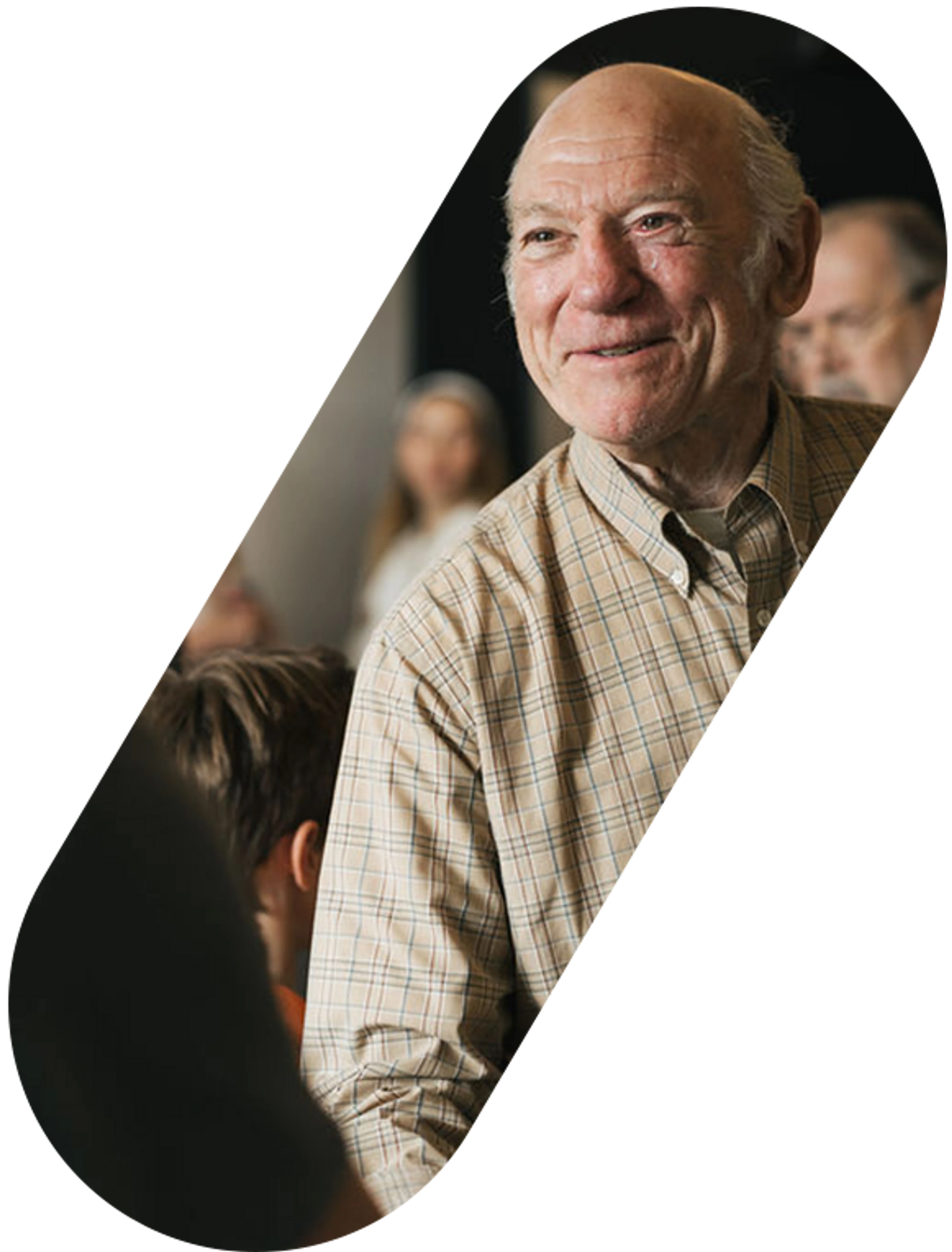 An elderly man with white hair, wearing a tan and blue plaid shirt, smiles while looking to his right. Other people are blurred in the background.