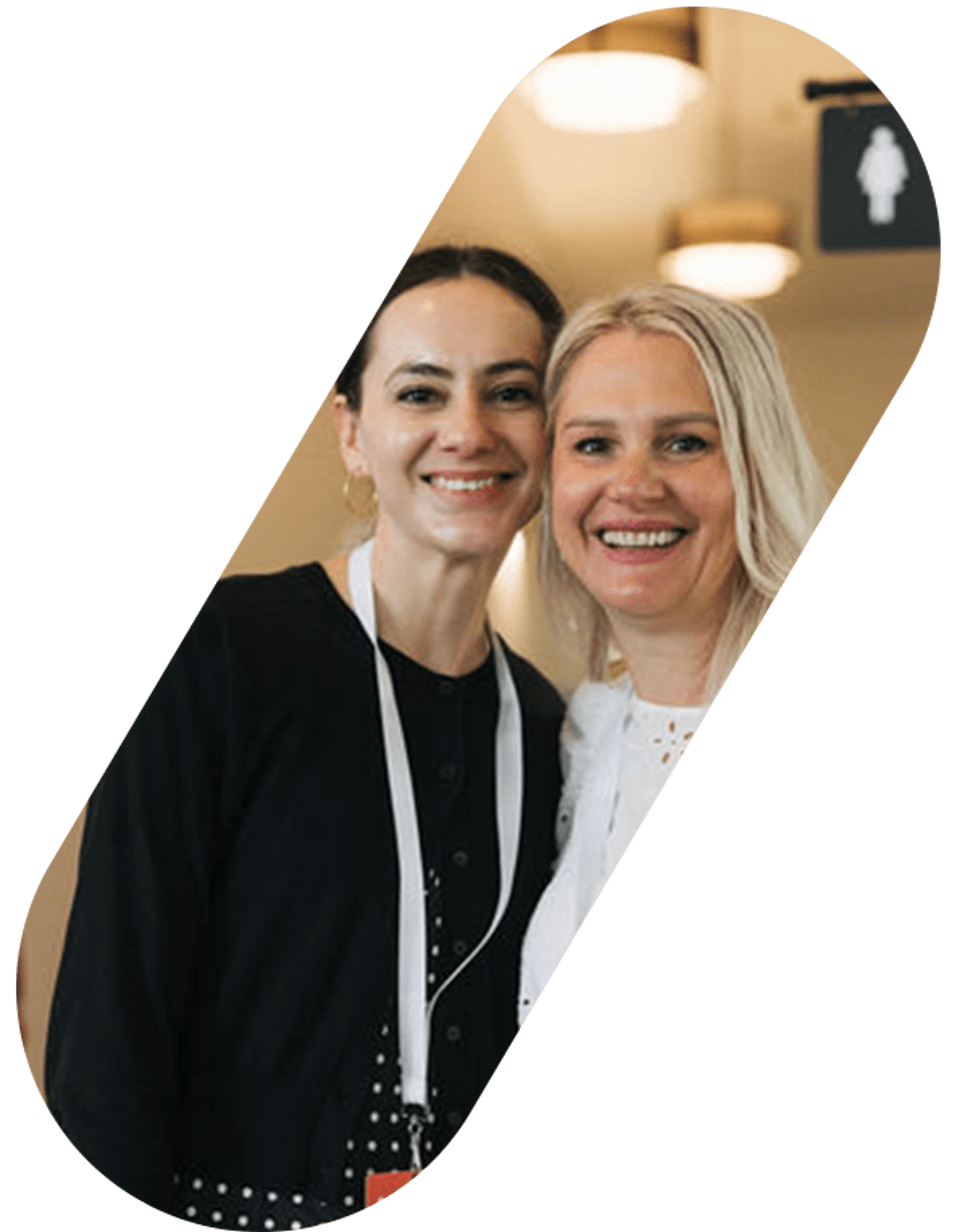 A close-up photo of two smiling women posing together. One has dark hair and wears a black top, while the other has blonde hair and wears a white top.