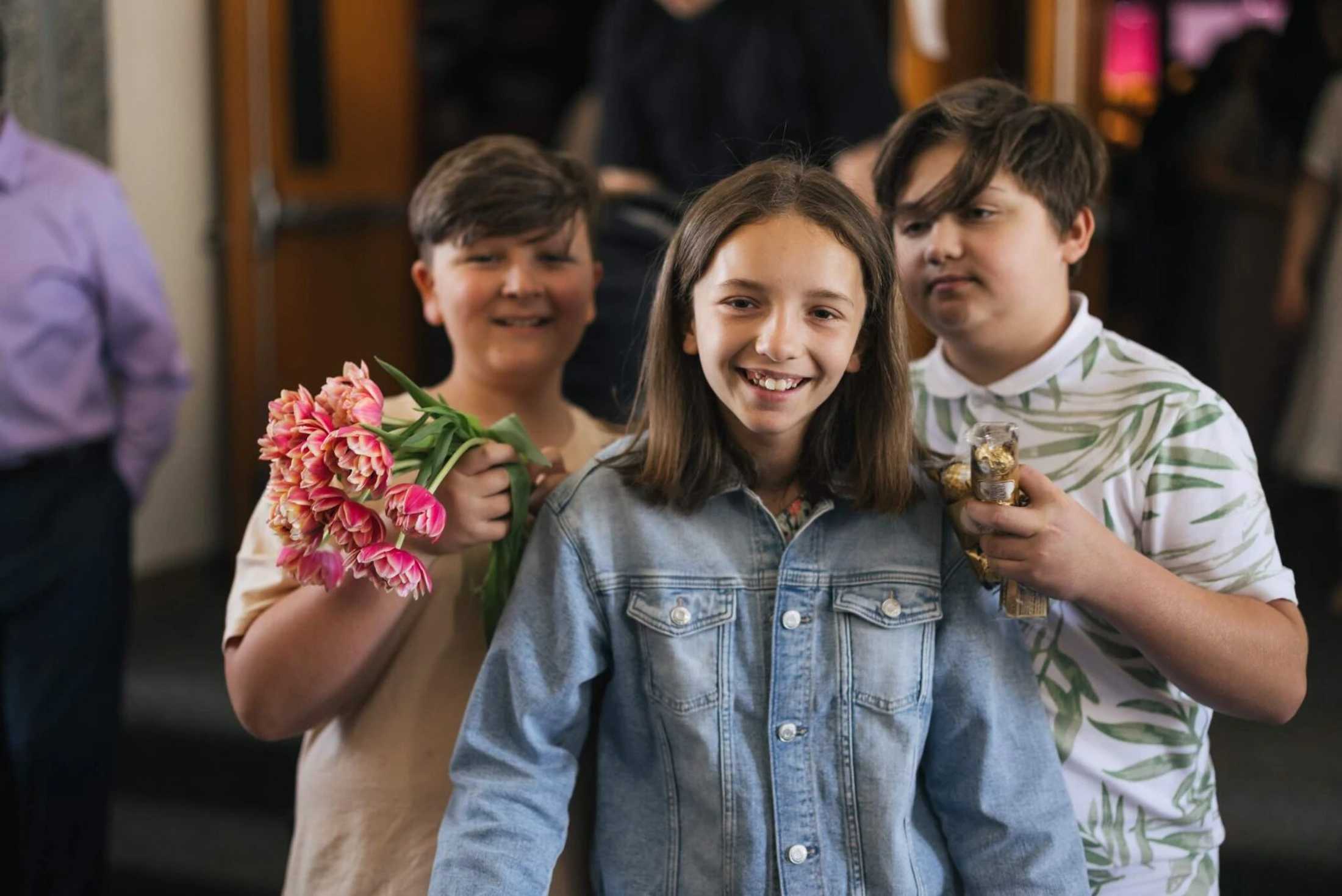 A girl in a denim jacket smiles between two boys. The boy on the left holds pink tulips and the boy on the right holds a stack of chocolates.
