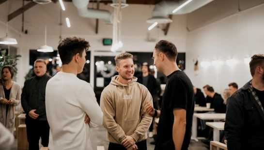 Three young men having a conversation in a well-lit room. The man in the center smiles warmly while wearing a beige hoodie with the word 'JESUS' on it.