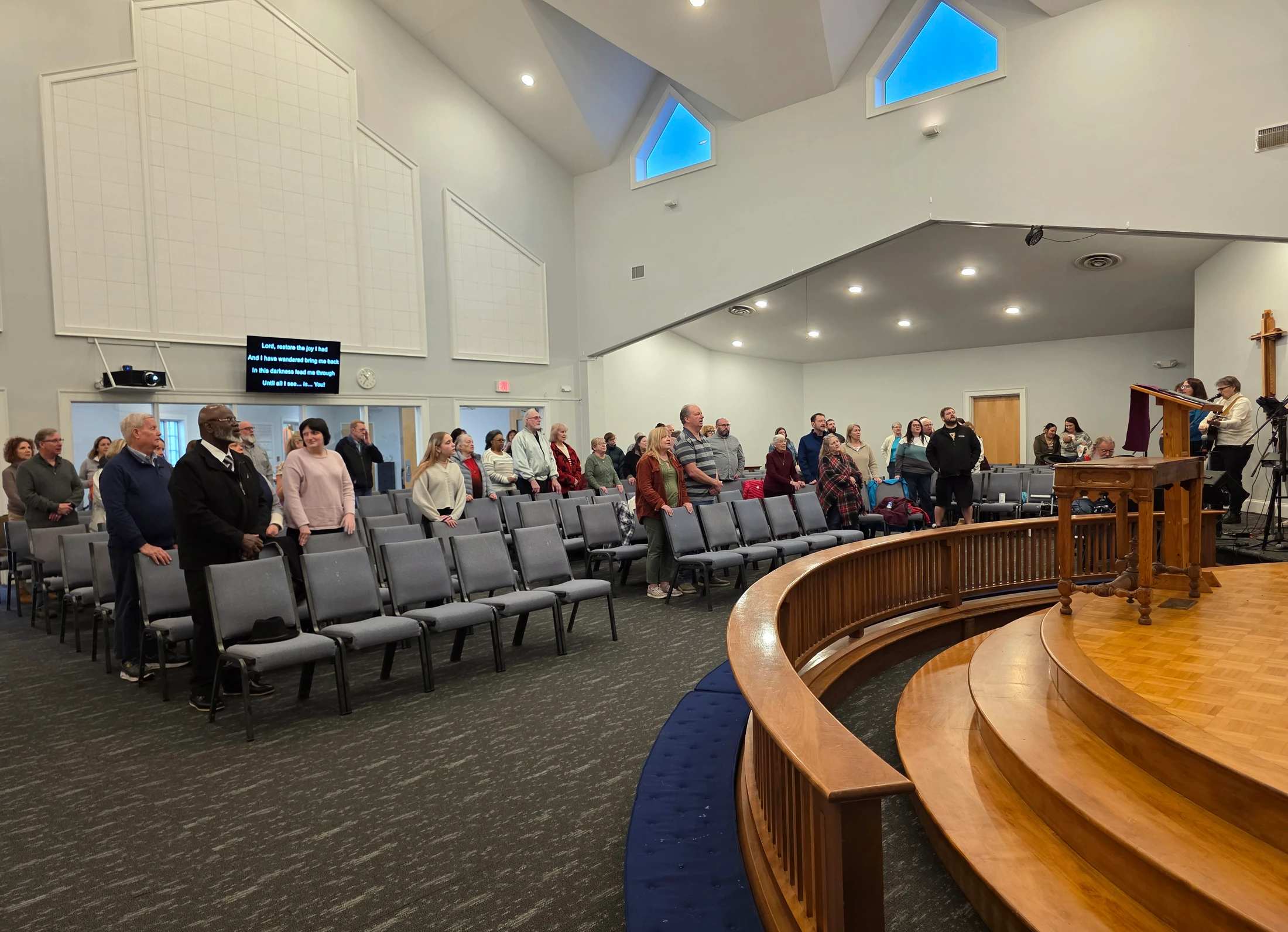 A diverse group of people stand in a modern church sanctuary during a service, singing along with lyrics displayed on a screen near the high ceiling.