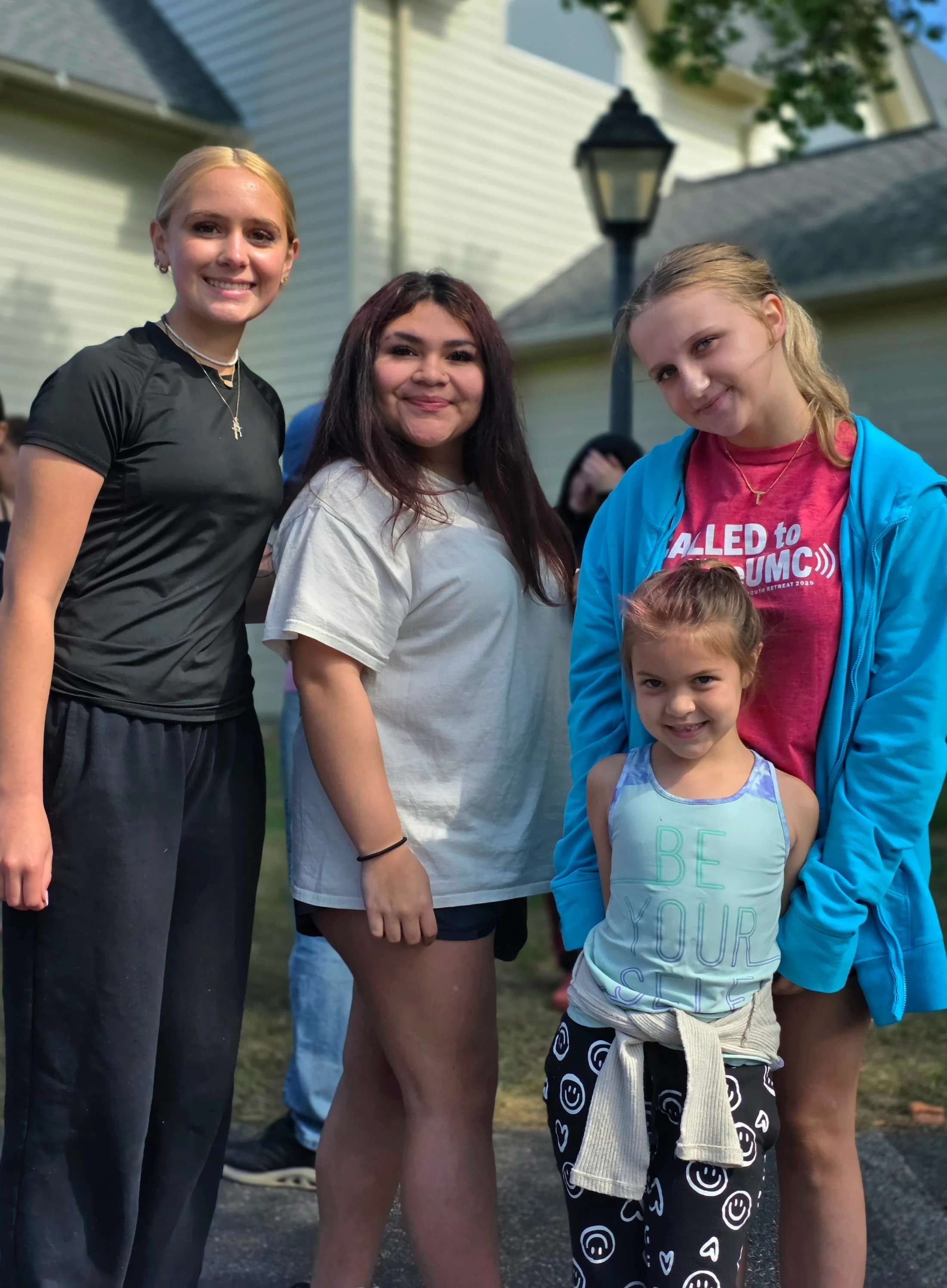 A group of three teenagers and one young girl stand together and smile for a photo outside in front of a house. They are all dressed in casual clothes.