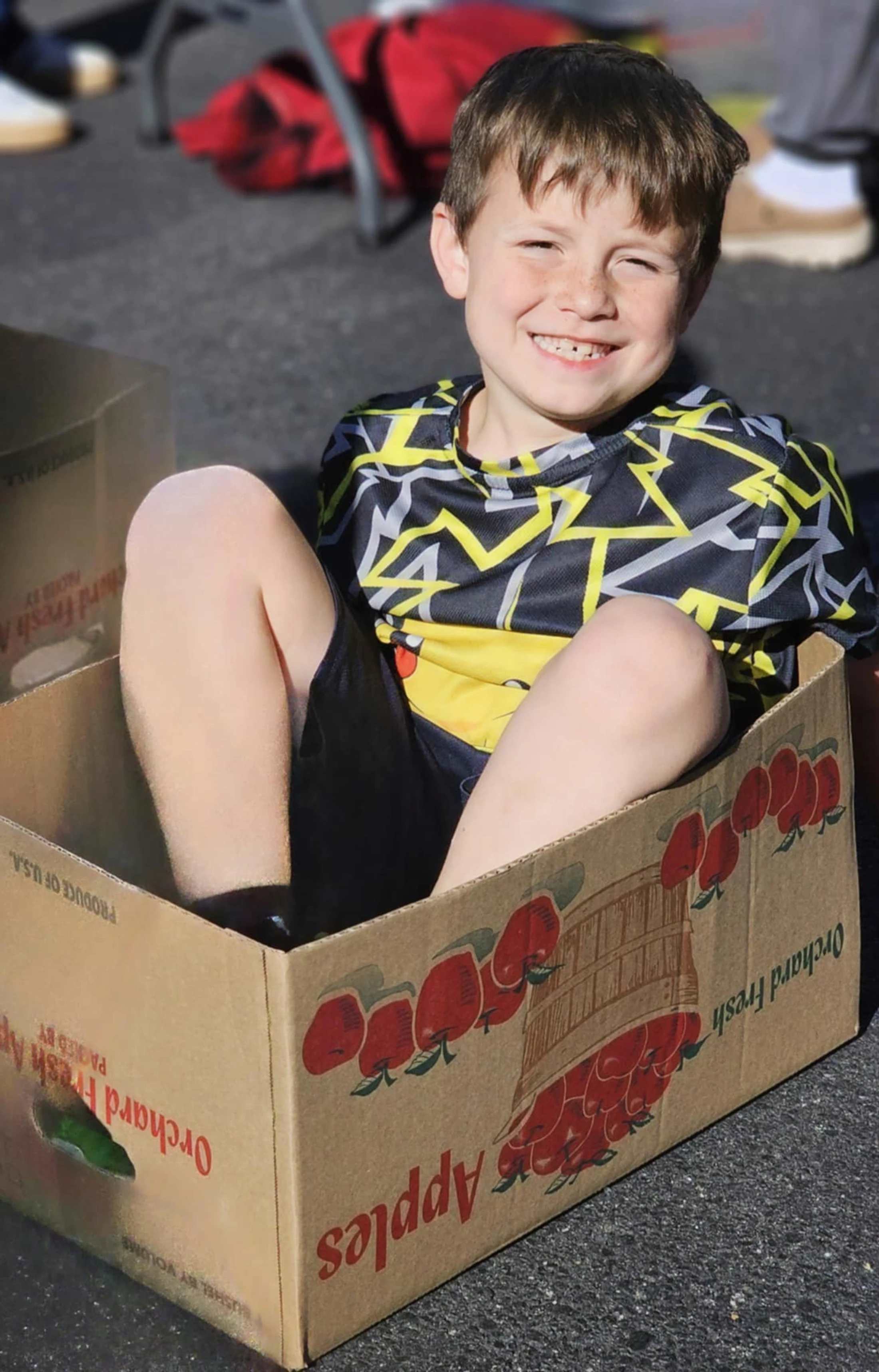 A smiling young boy with freckles sits inside a cardboard box for 'Orchard Fresh Apples' on a paved surface, looking happily at the camera.