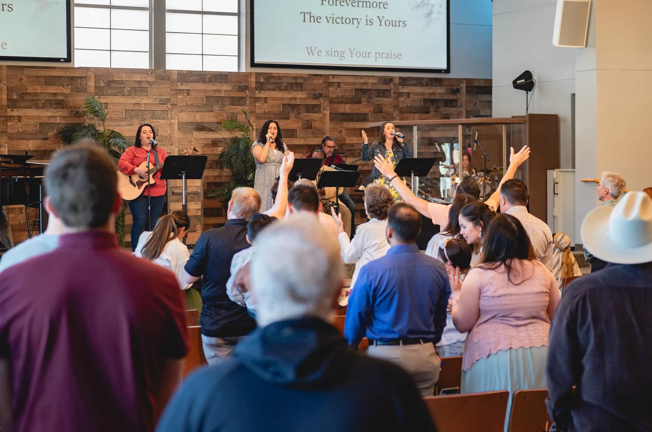 A church congregation, seen from behind, stands with hands raised in worship, facing a band singing and playing music on a stage with a wooden backdrop.