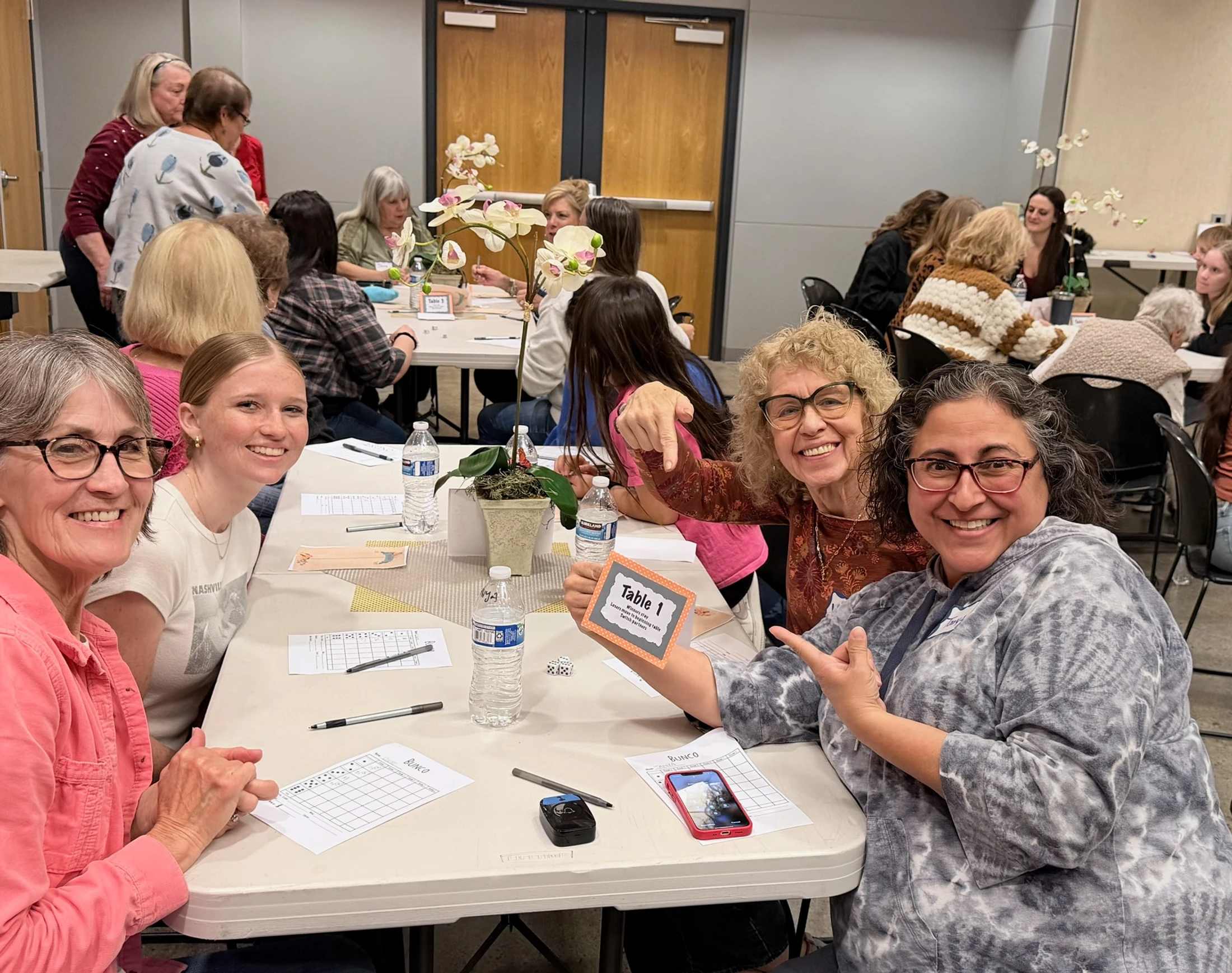 A group of women smile for the camera while sitting at a table during a Bunco game night. One woman holds up a sign that says 'Table 1'.