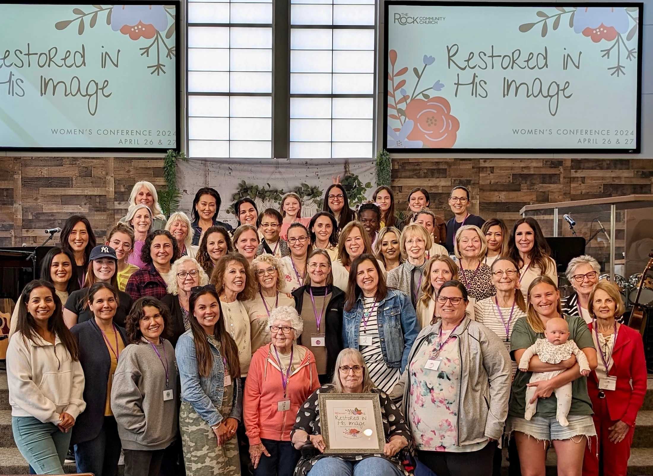 A large, diverse group of women smile for a photo inside a church during a conference titled 'Restored in His Image' displayed on screens behind them.