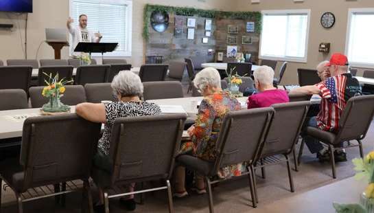 A man stands at a lectern speaking to a seated audience of mostly older adults in a community hall. The audience listens from rows of chairs.