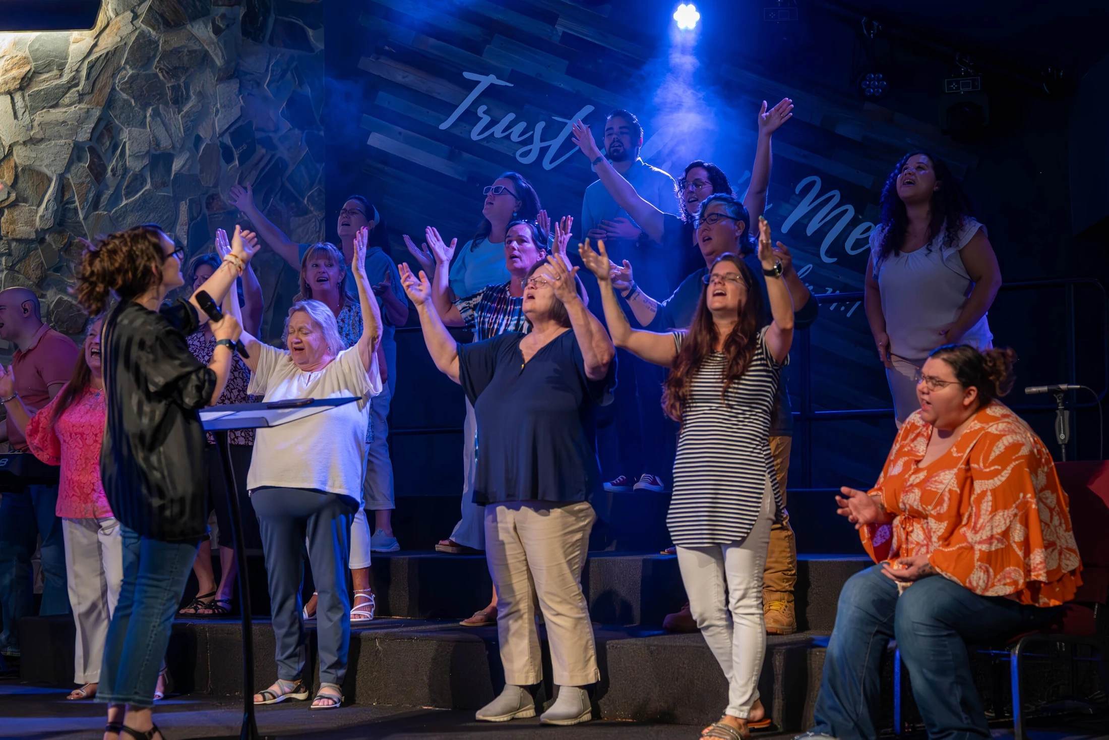 A diverse choir on a stage with blue lighting sings passionately with their hands raised in worship, while one woman leads them with a microphone.