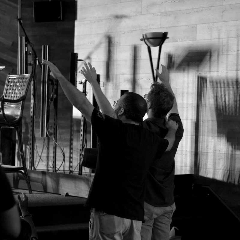 A black and white photo of two men from behind, standing with arms raised in worship. One man has his arm around the other's shoulder in a supportive gesture.