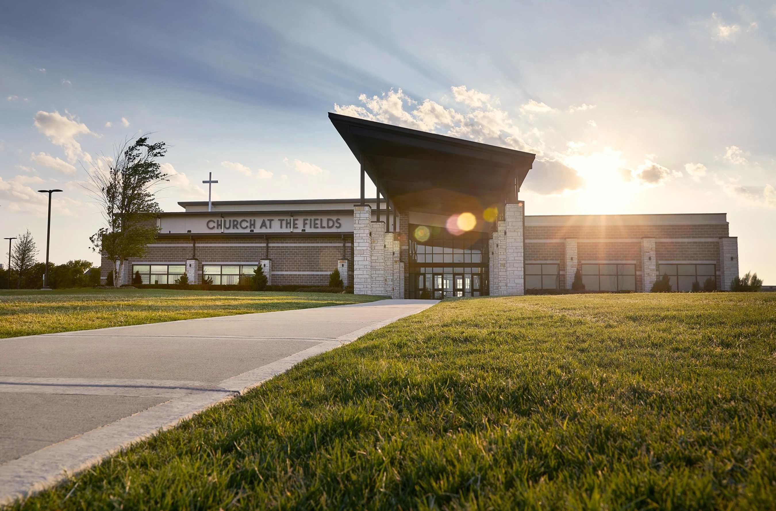 A low-angle exterior shot of the Church at the Fields building, with a walkway leading to the entrance as the sun sets in the background, creating lens flare.