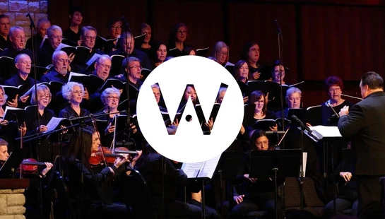 A wide view of a choir and orchestra performing on a stage with a stone wall background, led by a conductor. A white logo with a 'W' is in the center.