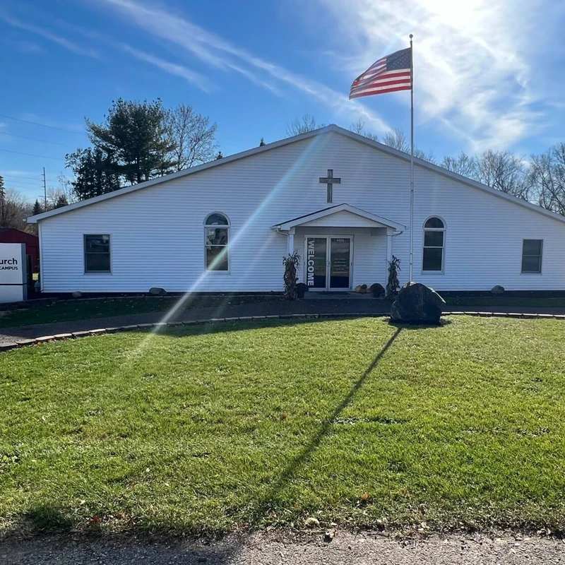 Exterior of a white, single-story church with a cross over the entrance. An American flag flies on a flagpole in the front lawn under a sunny, blue sky.