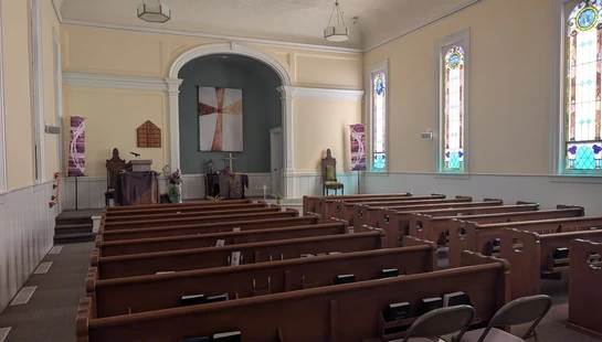 The empty interior of a sunlit church sanctuary, showing rows of wooden pews facing an altar with a pulpit and a cross tapestry under an archway.