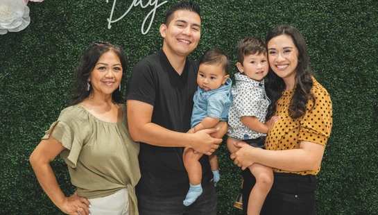 A family of five, including a grandmother, parents, and two young boys, smile for a photo in front of a green wall decorated with paper flowers.