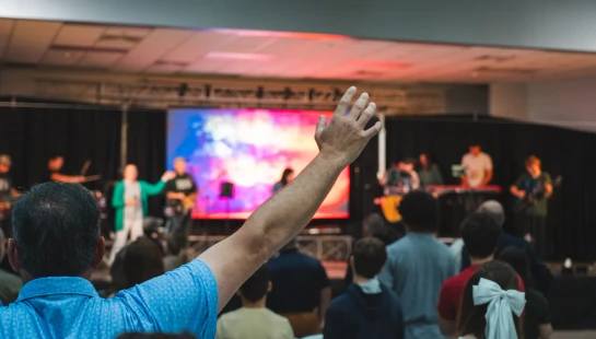 View from behind a man in a blue shirt with his arm raised, part of a crowd watching a band perform on a colorfully lit stage during a church service.