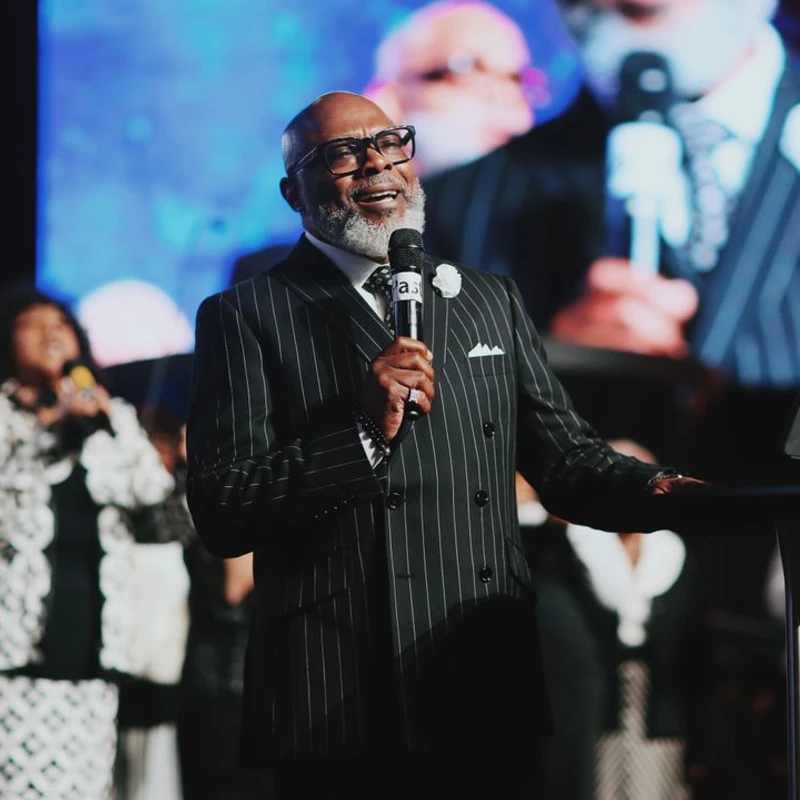 An older Black man with a gray beard and glasses, wearing a pinstripe suit, smiles while speaking into a microphone on stage in front of a choir.